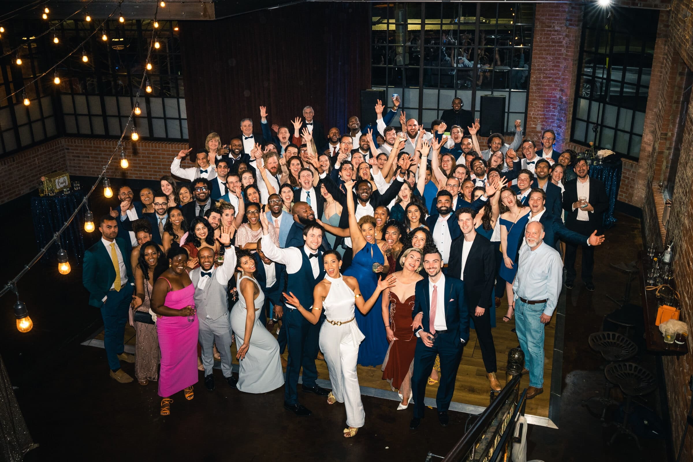 duke chapel & cookery wedding - sasha & ben A large group of people dressed in formal attire poses together in a spacious room adorned with string lights. Smiling and raising their hands in celebration, they are reminiscent of gatherings at Duke Chapel, surrounded by charming brick walls and large windows.