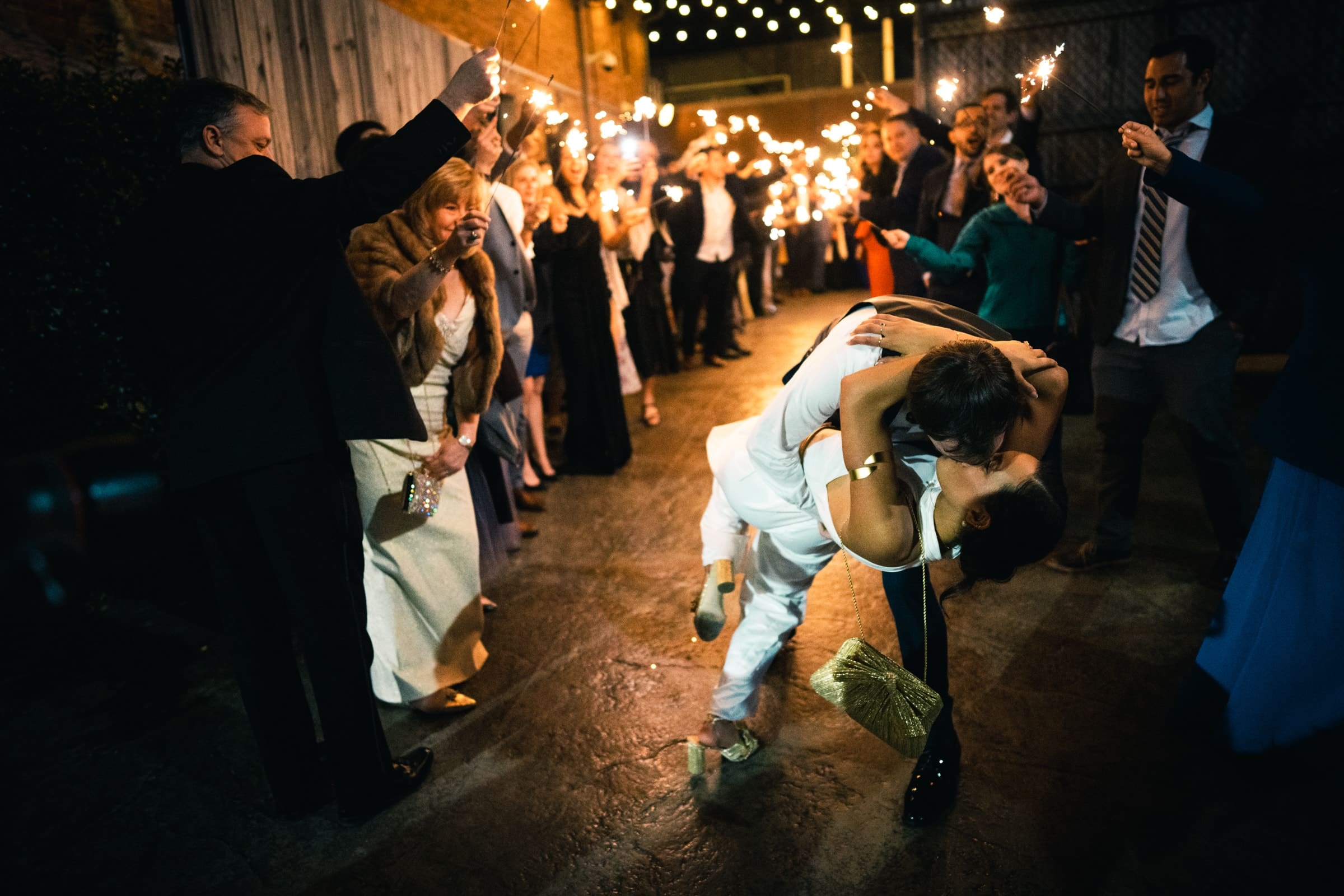 duke chapel & cookery wedding - sasha & ben A couple shares a romantic dip kiss on a dimly lit path near Duke Chapel, surrounded by cheering friends holding sparklers. The festive atmosphere is complemented by string lights overhead, creating a warm, celebratory scene.
