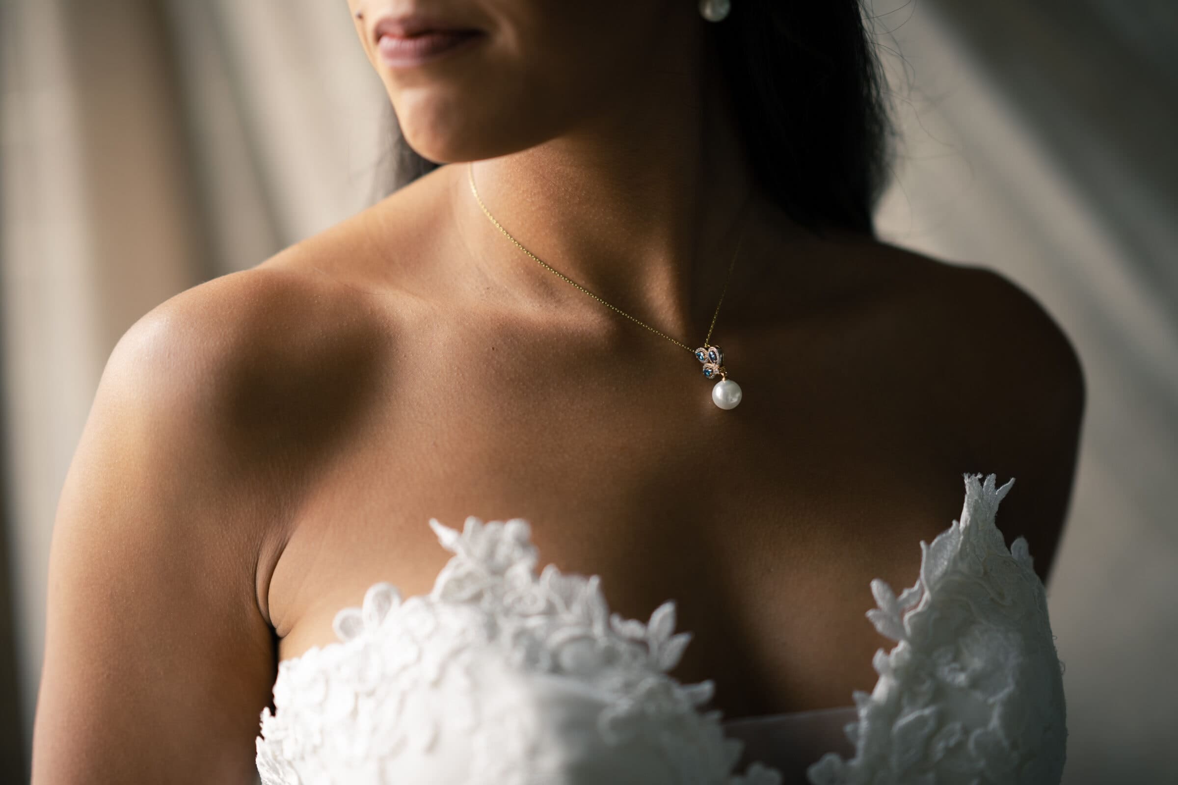 duke chapel & cookery wedding - sasha & ben In the gentle ambiance, a woman stands in a white lace strapless dress and pearl necklace, subtly reminiscent of the regal beauty of Duke Chapel. Her dark hair and light makeup enhance the serene mood as she faces slightly to the side.