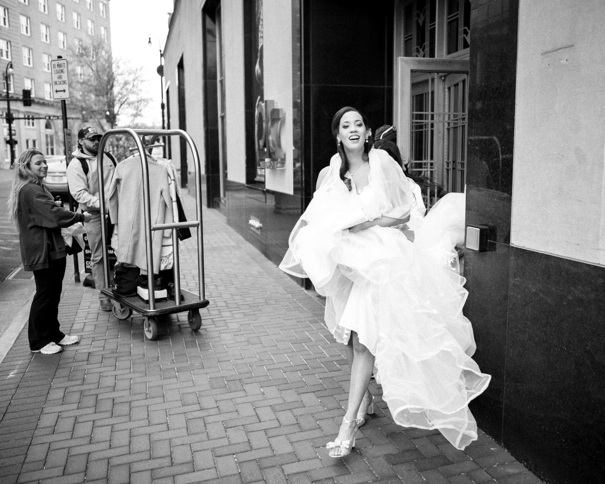 duke chapel & cookery wedding - sasha & ben A bride joyfully exits Duke Chapel, holding her wedding dress above her ankles. On the sidewalk, people pass by, including someone pushing a luggage cart. The urban street setting is captured beautifully in black and white.