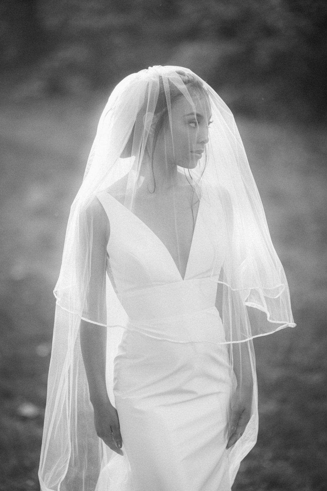 A woman in a wedding dress stands outdoors at an Umstead weddings venue, her veil partially covering her face. The dress features a deep V-neckline. The black-and-white image captures a soft, timeless effect with blurred trees in the background, exemplifying Umstead wedding photography.