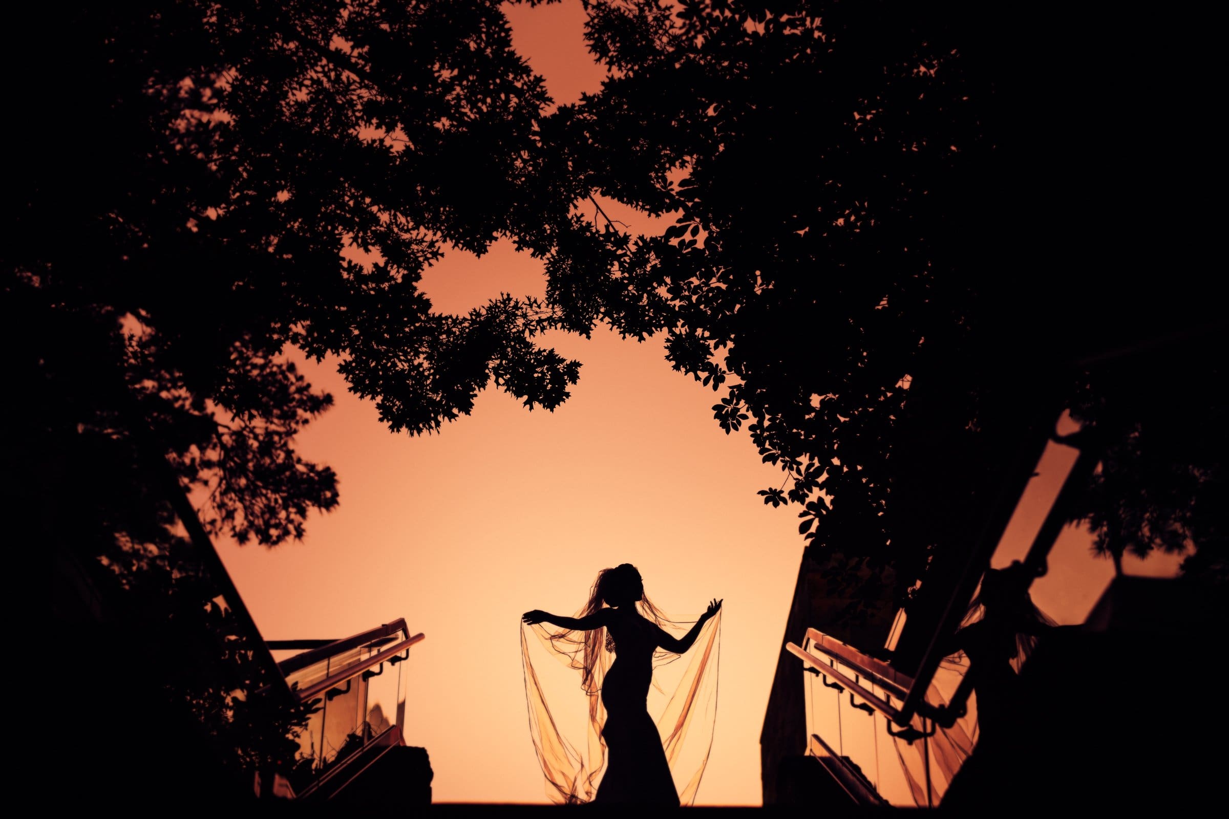 Silhouette of a person holding a flowing fabric, standing between trees and escalators, with an orange sky in the background—a scene reminiscent of the enchanting elegance found in Umstead wedding photography.