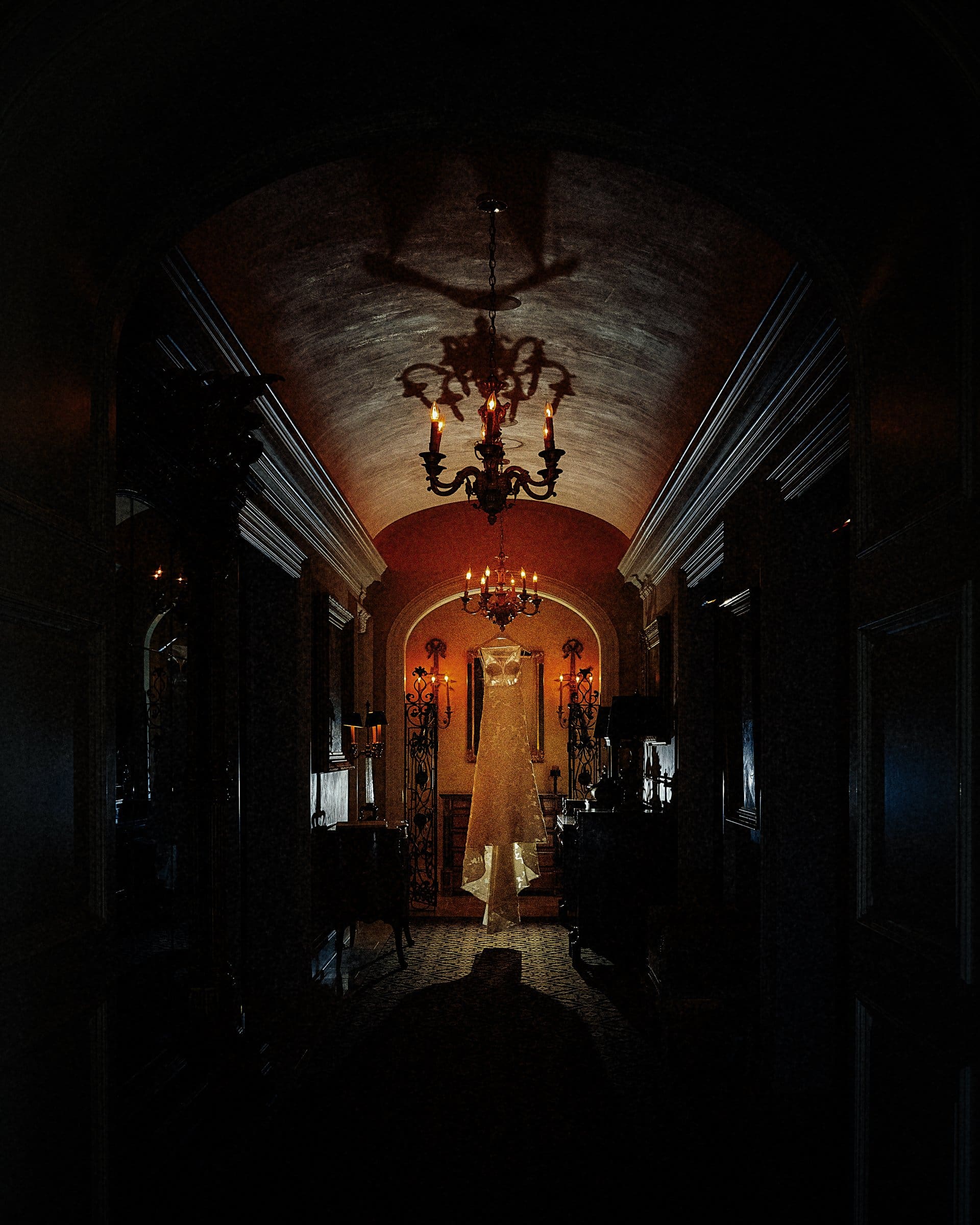 A wedding dress is dramatically illuminated, hanging in a dimly lit, ornate hallway. Captured by a Raleigh photographer, an elegant chandelier casts shadows on the curved ceiling, enhancing the room's vintage ambience.