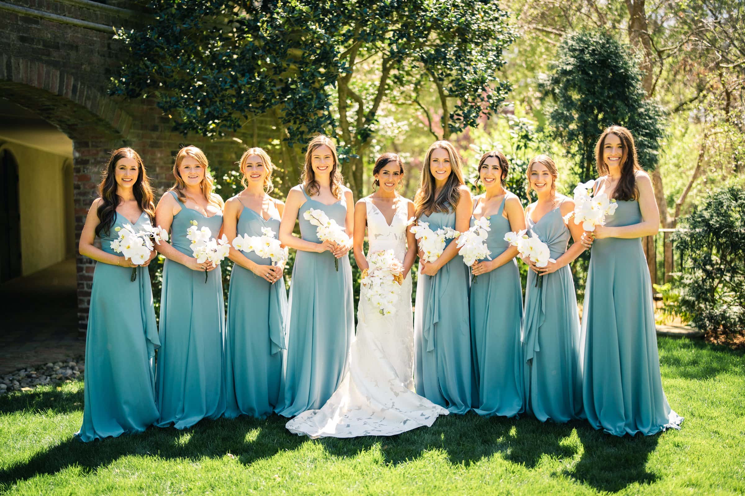 A bride stands outdoors in a garden with eight bridesmaids in matching blue dresses, captured perfectly by a Raleigh photographer. Each holds a bouquet of white flowers. Sunlight filters through the trees, casting dappled shadows on the grass. They are all smiling and looking at the camera.