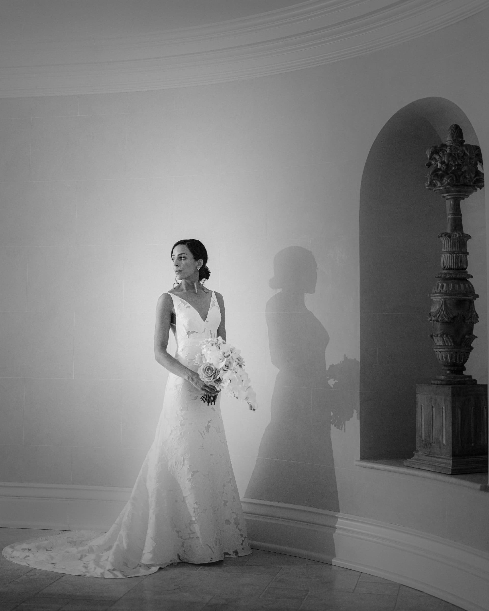 A bride in a long white gown holds a bouquet, captured beautifully by a Raleigh photographer as she looks to her left, casting a shadow on the curved wall. The setting is simple, with a decorative column in a nook. The image is elegantly rendered in black and white.