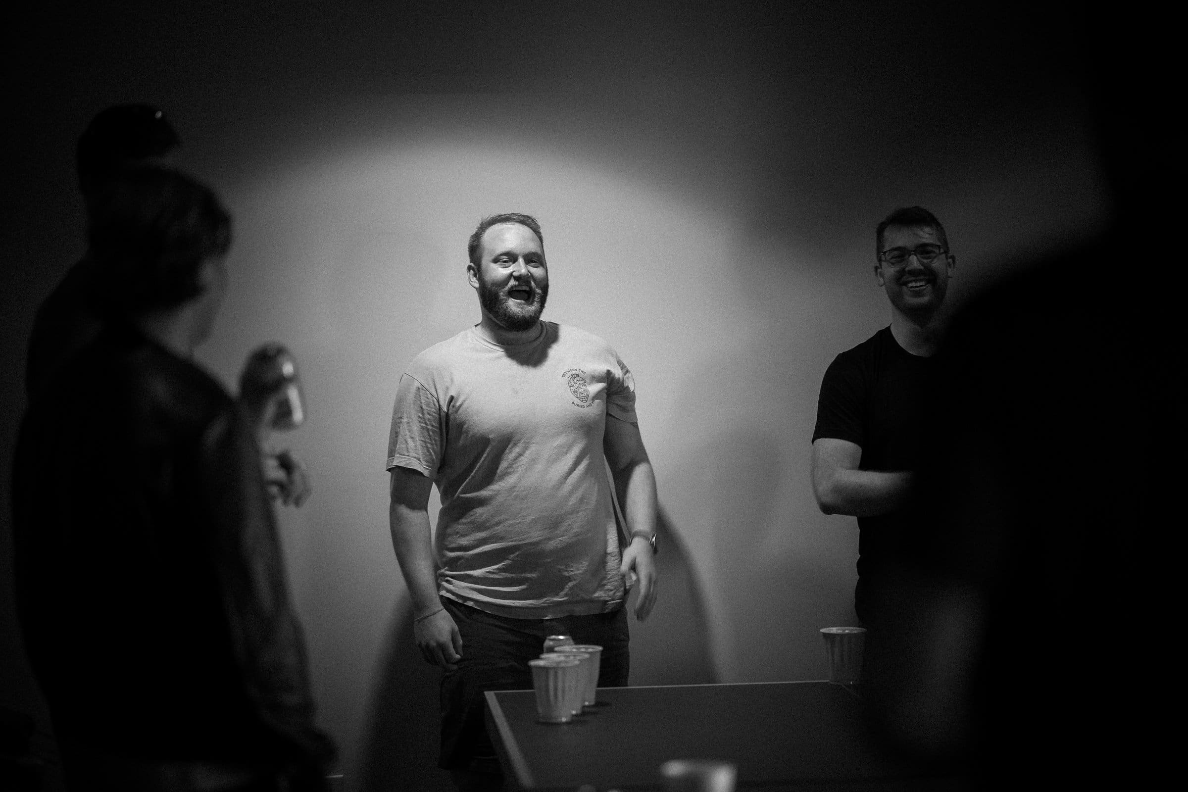 A group gathers around a table in a dimly lit room, smiles all around as they hold drinks. Captured by a Raleigh photographer, the spotlight falls on two men at the center—one in a light t-shirt and the other in dark attire—with cups scattered on the table before them.