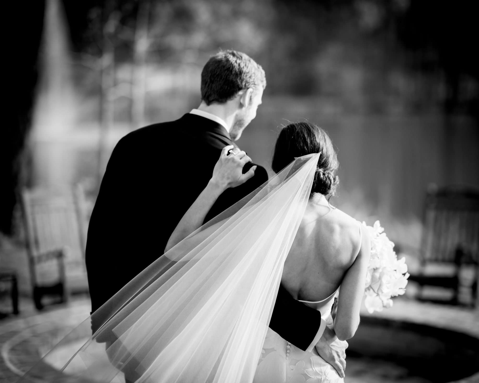 Captured by a Raleigh photographer, this black and white photo beautifully illustrates a newlywed couple walking away, the groom’s arm lovingly around the bride. Her bouquet and flowing veil add elegance against the blurred backdrop of trees and chairs in this outdoor setting.