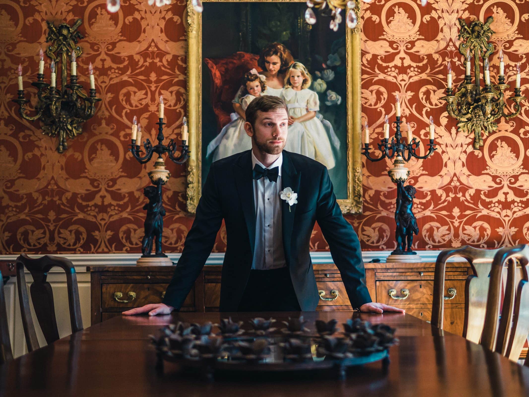 A Raleigh photographer captures a man in a tuxedo standing at the end of a dining table in an ornate room, adorned with red patterned wallpaper, vintage chandeliers, and a large painting of two children above a wooden sideboard.