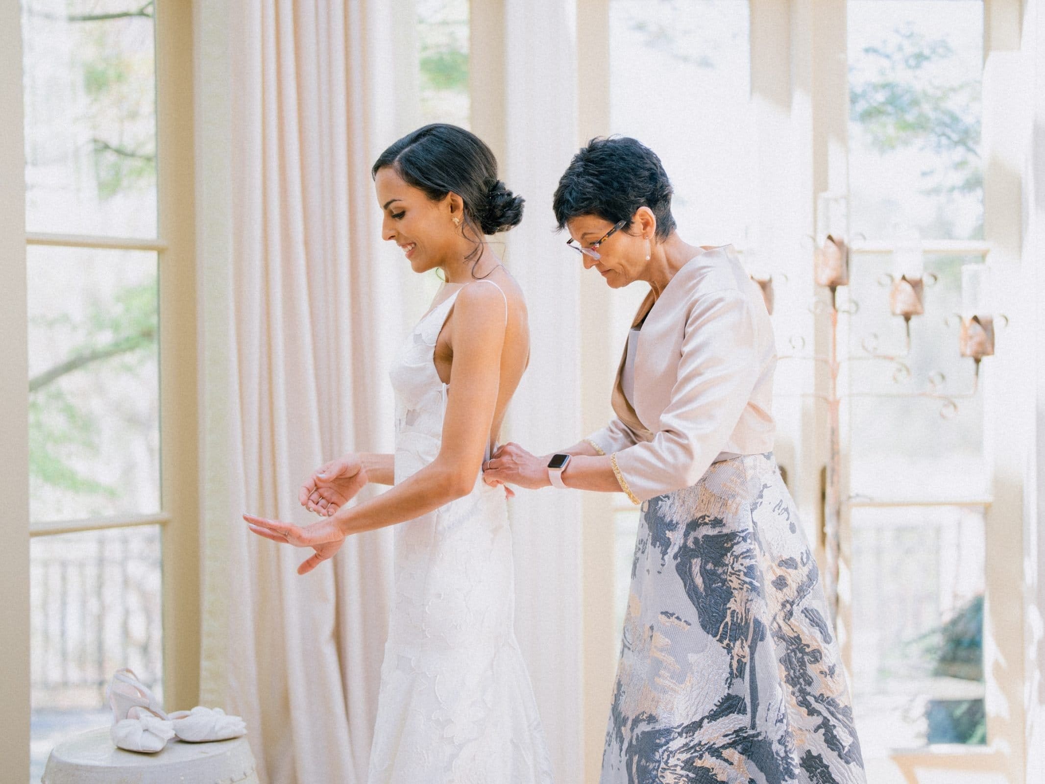 A bride in a white gown smiles as a woman in an elegant floral dress helps with the fitting. Captured by a Raleigh photographer, they stand in a bright room with large windows and natural light. Nearby, a pair of white shoes rests on a small table.