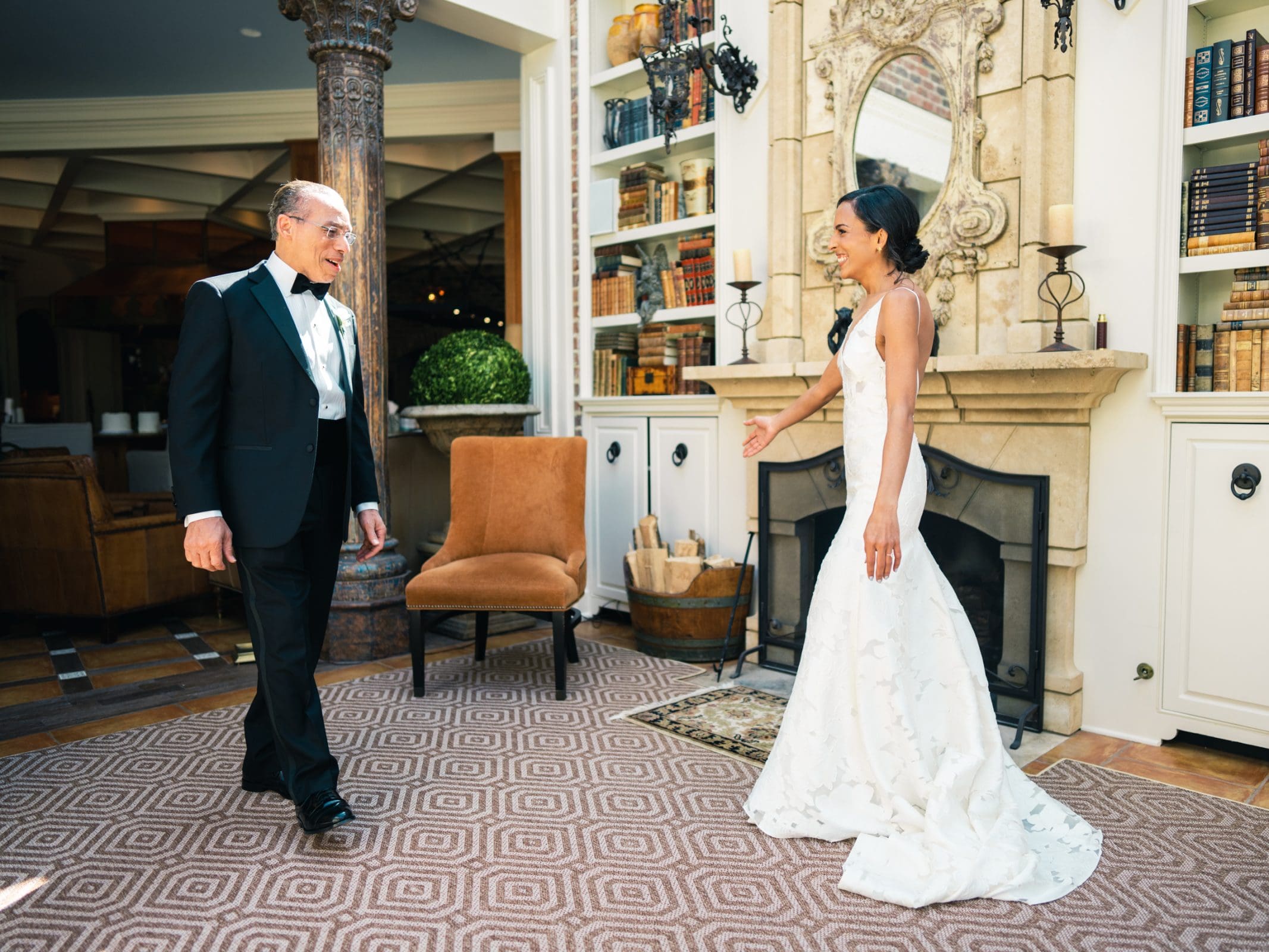 A Raleigh photographer captures the joyful bride in a white gown as she approaches a man in a tuxedo inside a warmly decorated room with bookshelves and a fireplace, sharing a happy moment.