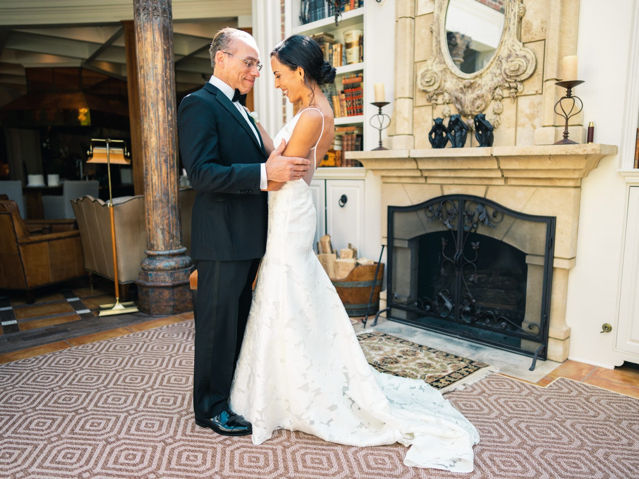 A couple in formal attire smiles at each other while dancing in an elegant room, beautifully captured by a Raleigh photographer. The woman wears a white gown and the man a black tuxedo near a fireplace with bookshelves and decorative columns in the background.