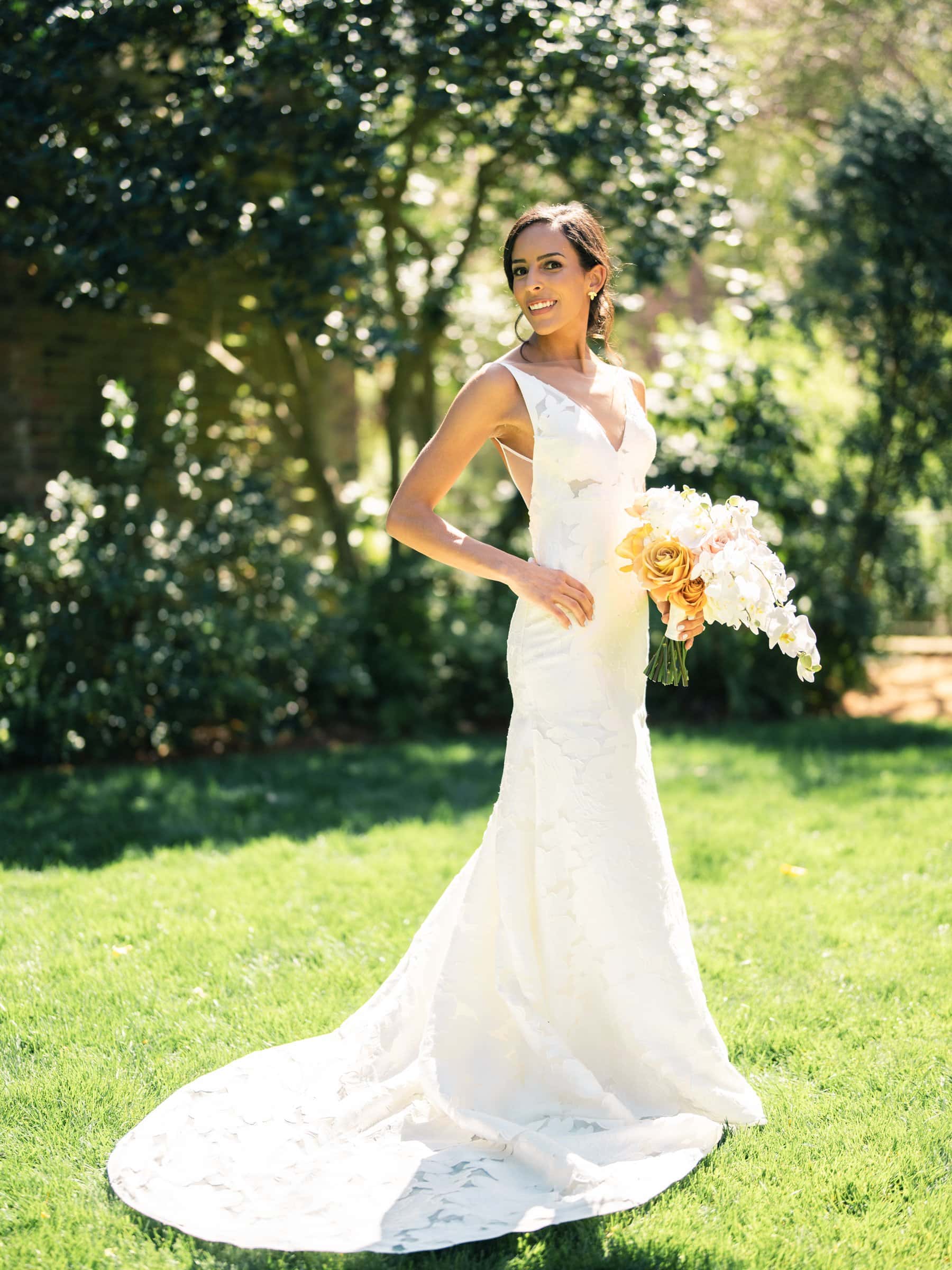 A bride stands outdoors on a sunny day, captured by a Raleigh photographer. She wears a white sleeveless wedding gown with a long train and holds a bouquet of yellow and white flowers. Behind her, trees with lush green foliage complete the picturesque scene.