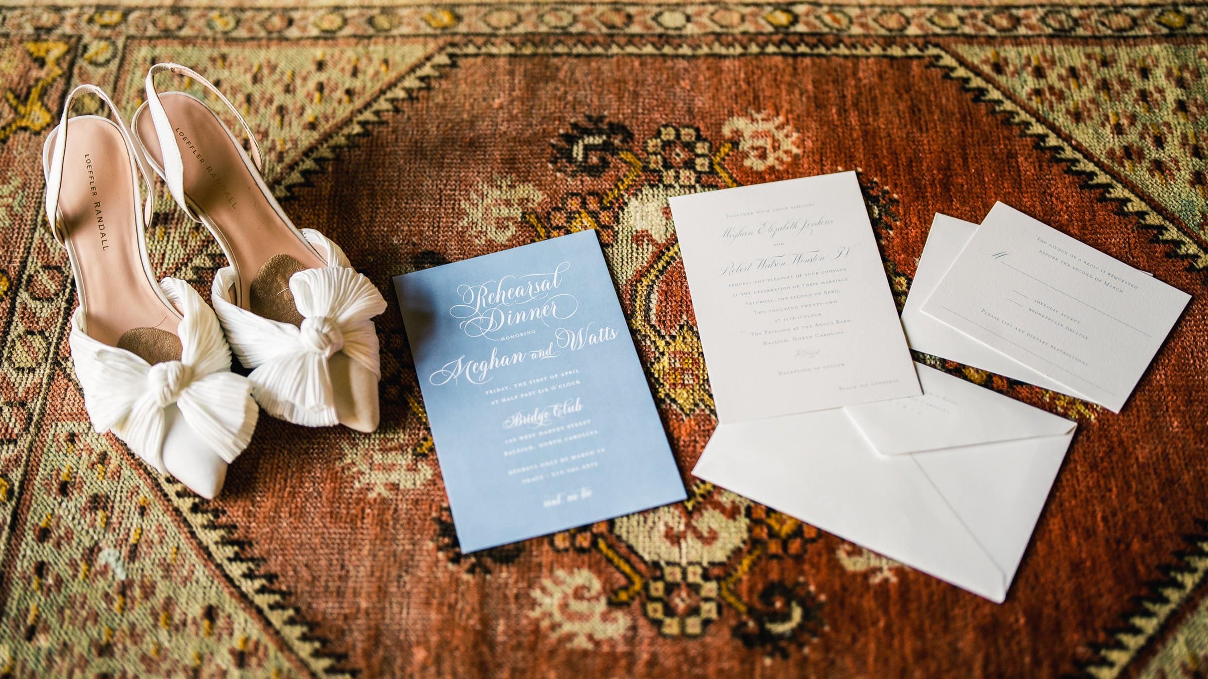 A pair of elegant white high heels with bows sits gracefully on a patterned rug, as though captured by a Raleigh photographer. Nearby, amidst the artistry, lie several stationery items: a blue rehearsal dinner invitation and crisp white envelopes.