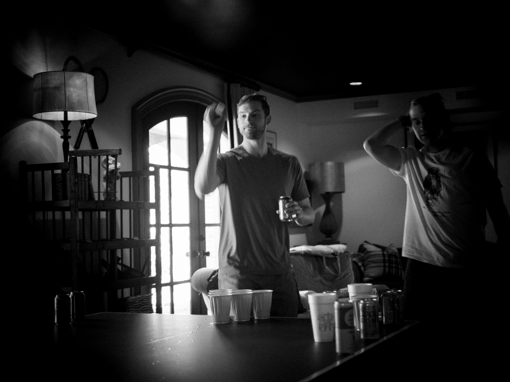 A black and white image captured by a Raleigh photographer showcases two people playing beer pong in a dimly lit room. One tosses a ball towards cups on the table, while the other stands nearby. A shelf, lamp, and closed door add depth in the background.