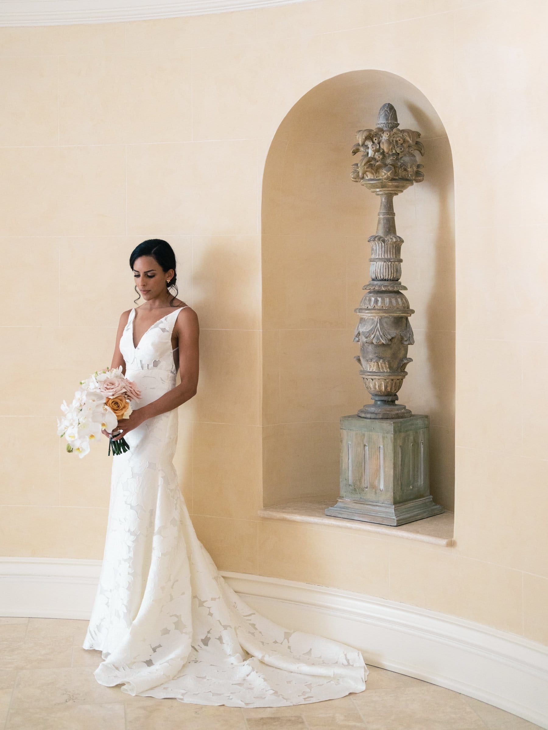 A bride in a white, sleeveless wedding gown and holding a bouquet stands next to an ornate sculpted pedestal in a softly lit alcove with a cream-colored wall, captured by a Raleigh photographer.