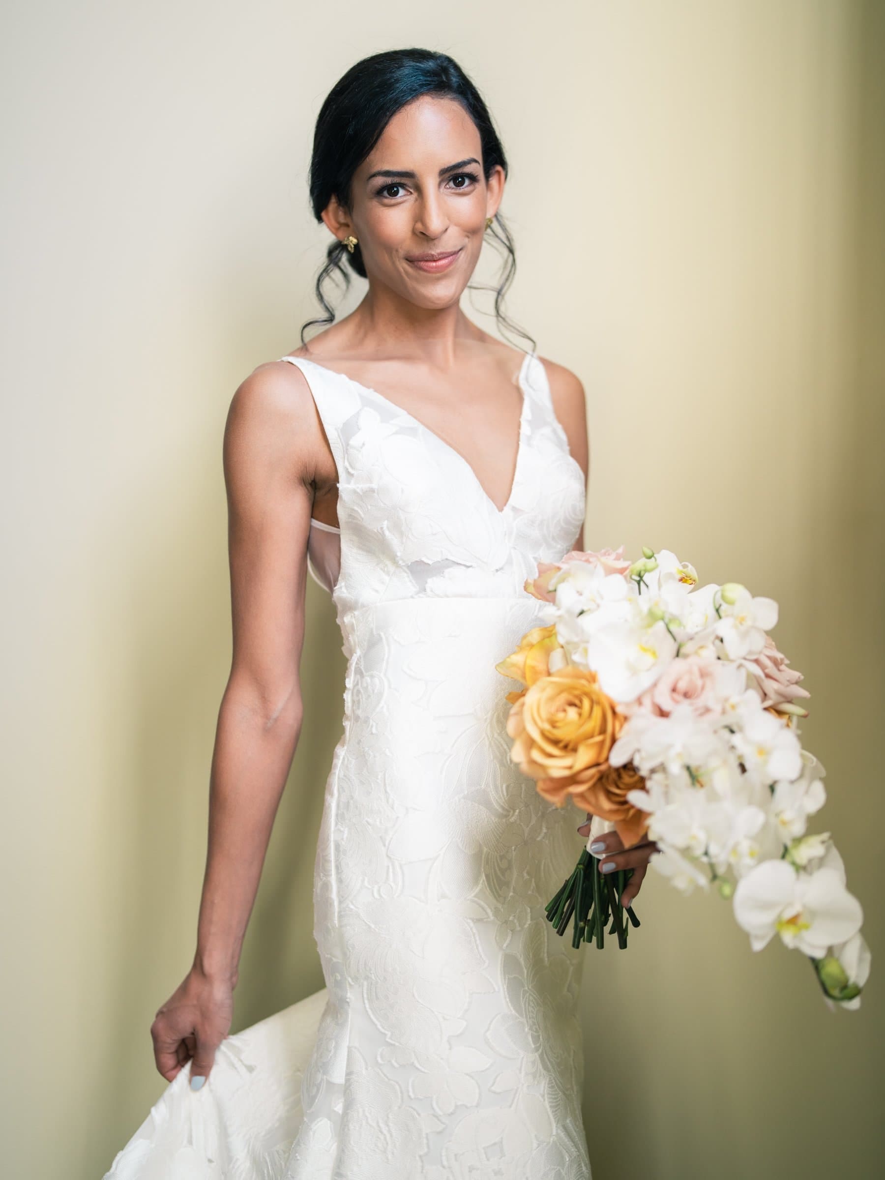 A bride in a sleeveless white gown, captured by a Raleigh photographer, holds a bouquet of yellow, pink, and white flowers. She stands against a light-colored background, smiling softly. Her hair is styled in a low bun with loose waves.