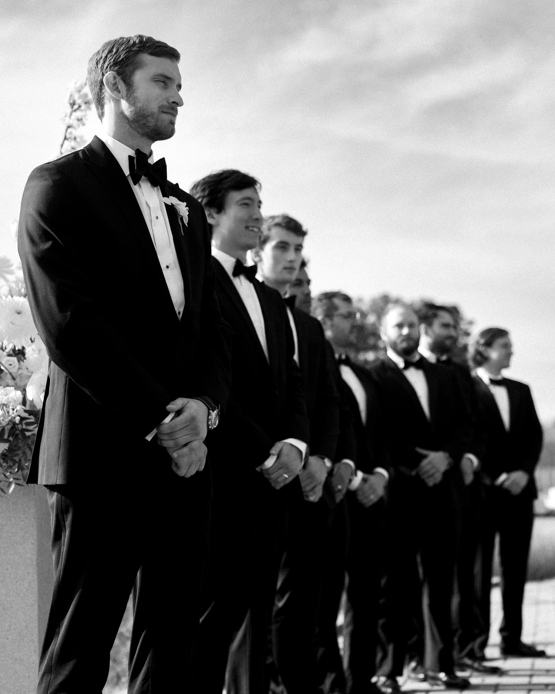 A group of groomsmen in tuxedos stand in a line outdoors, looking forward. Some wear bow ties. The background, captured beautifully by a Raleigh photographer, shows a clear sky and floral arrangements, suggesting a formal event or wedding ceremony.