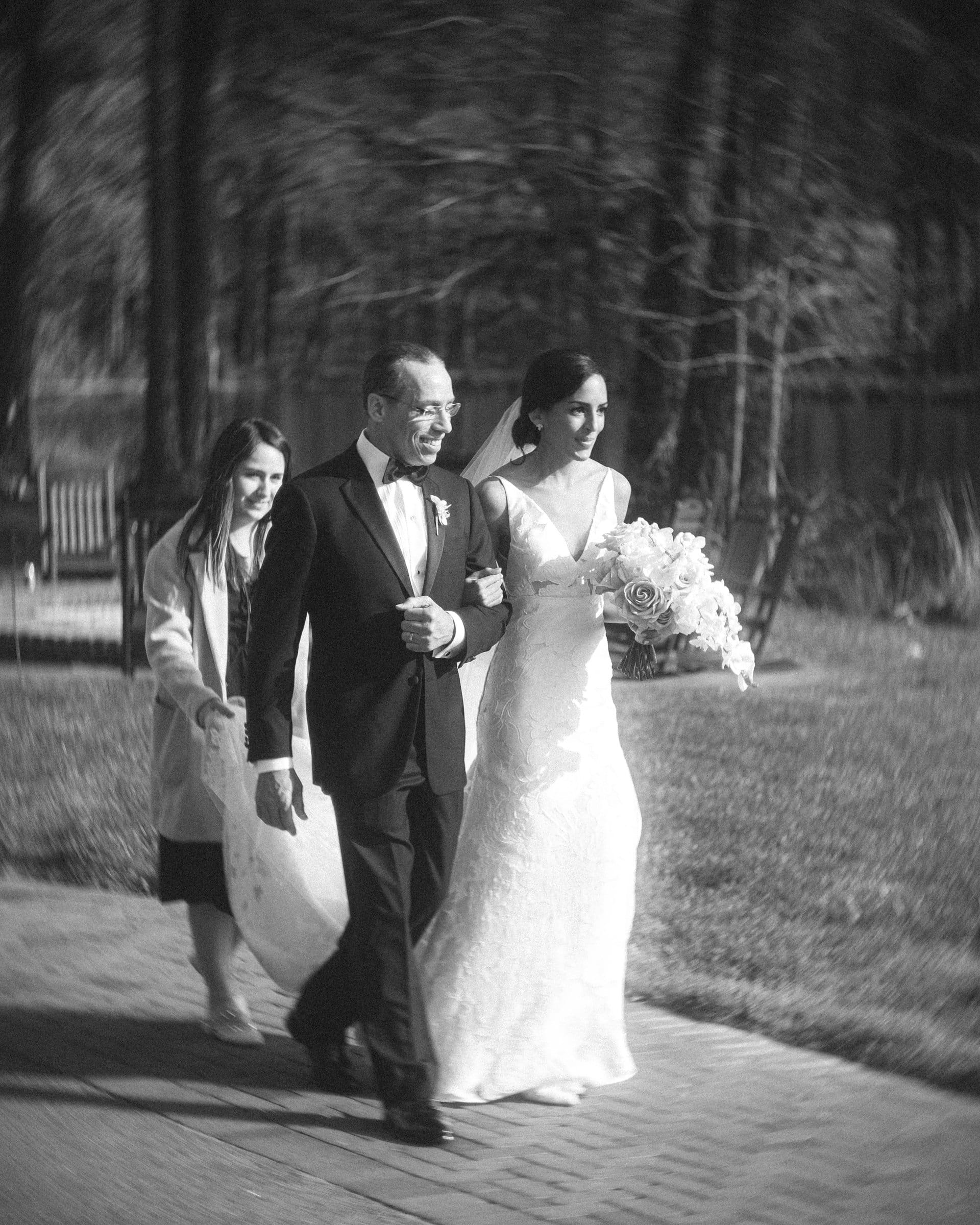 A black and white photo, captured by a Raleigh photographer, shows a bride in a wedding dress walking arm-in-arm with an older man in a tuxedo. A young girl follows, holding the train of the bride's dress. They are outdoors on a path, with trees and benches in the background.
