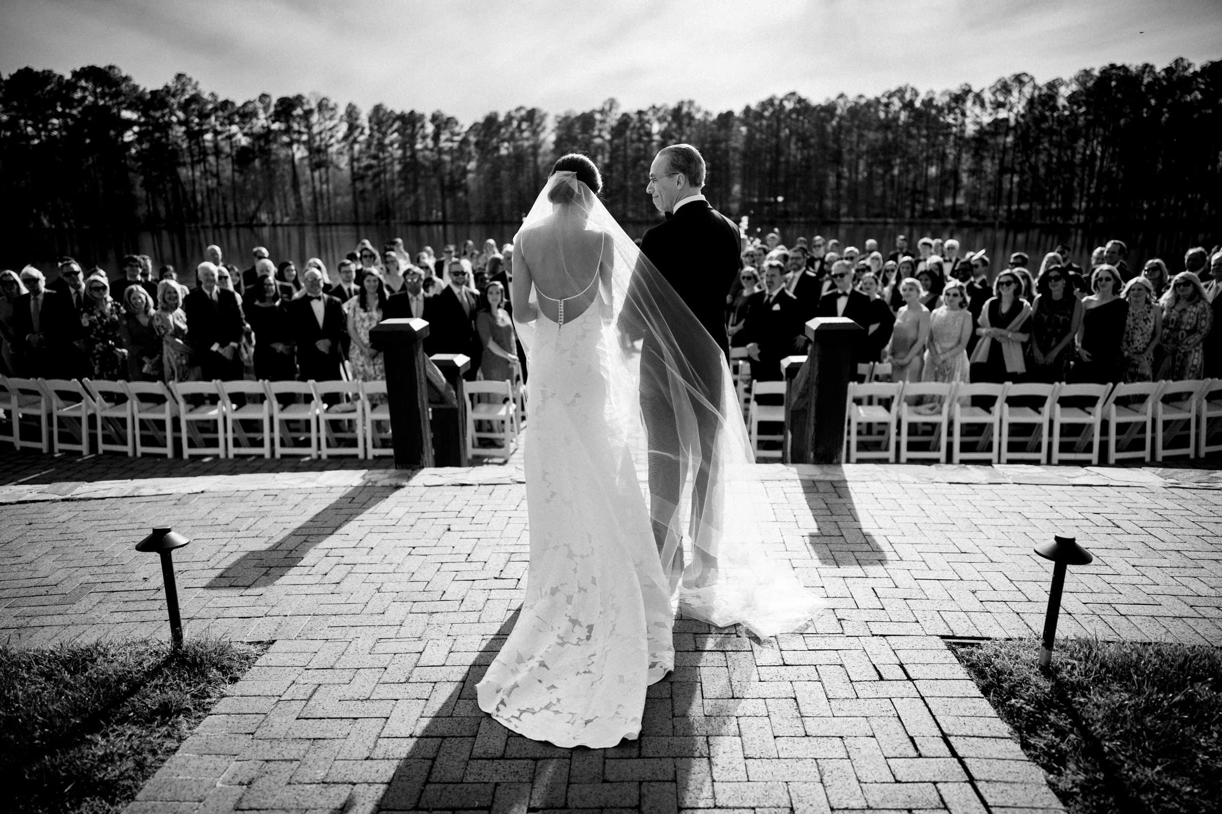 A bride and groom stand before a seated crowd during an outdoor wedding ceremony, perfectly captured by a Raleigh photographer. The bride's veil flows behind her on the brick pathway, with trees visible in the background under a clear sky.