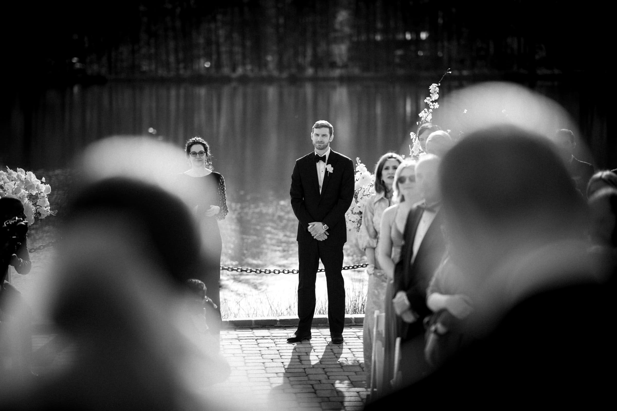 Captured by a Raleigh photographer, this black and white image portrays a groom standing at an outdoor lakeside wedding ceremony. Clad in a suit and bowtie, he gazes down the aisle as guests blur softly in the foreground.