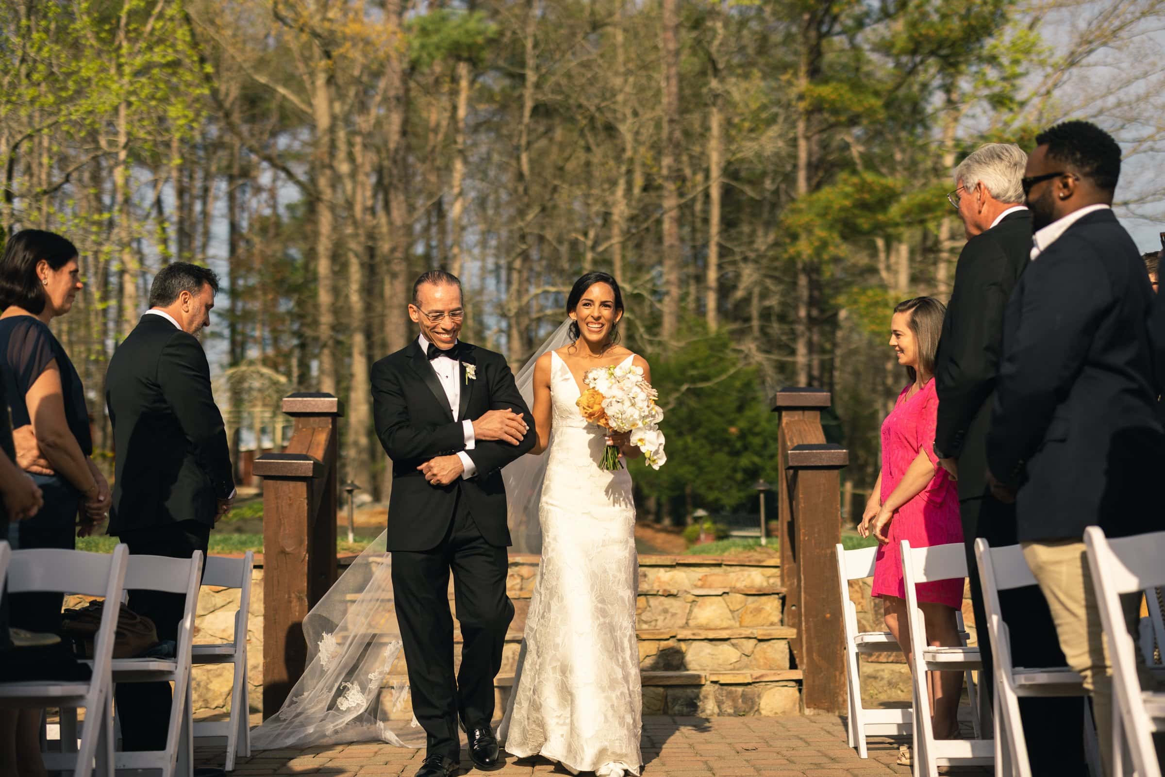 A bride in a white dress holding a bouquet and an older man in a suit walk down the outdoor aisle, captured beautifully by a Raleigh photographer. Guests sit on either side, observing. Behind them lies a stone staircase and a lush forest backdrop under a clear sky.