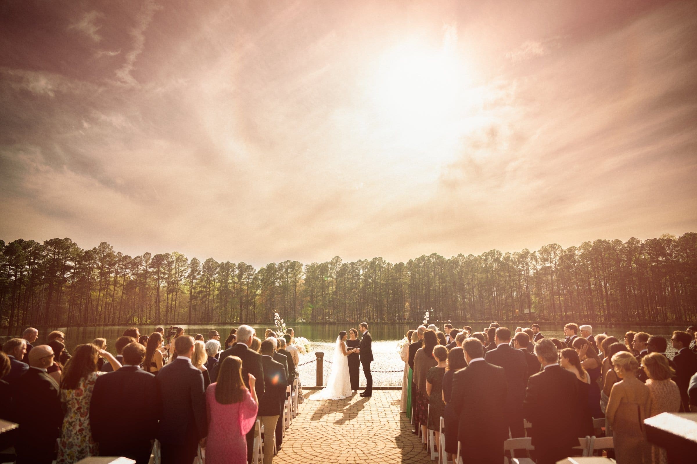 A couple stands together, exchanging vows under a bright sun at an outdoor wedding ceremony, captured by a Raleigh photographer. Surrounded by seated guests, the background features tall trees and a clear sky, creating a serene and picturesque setting.