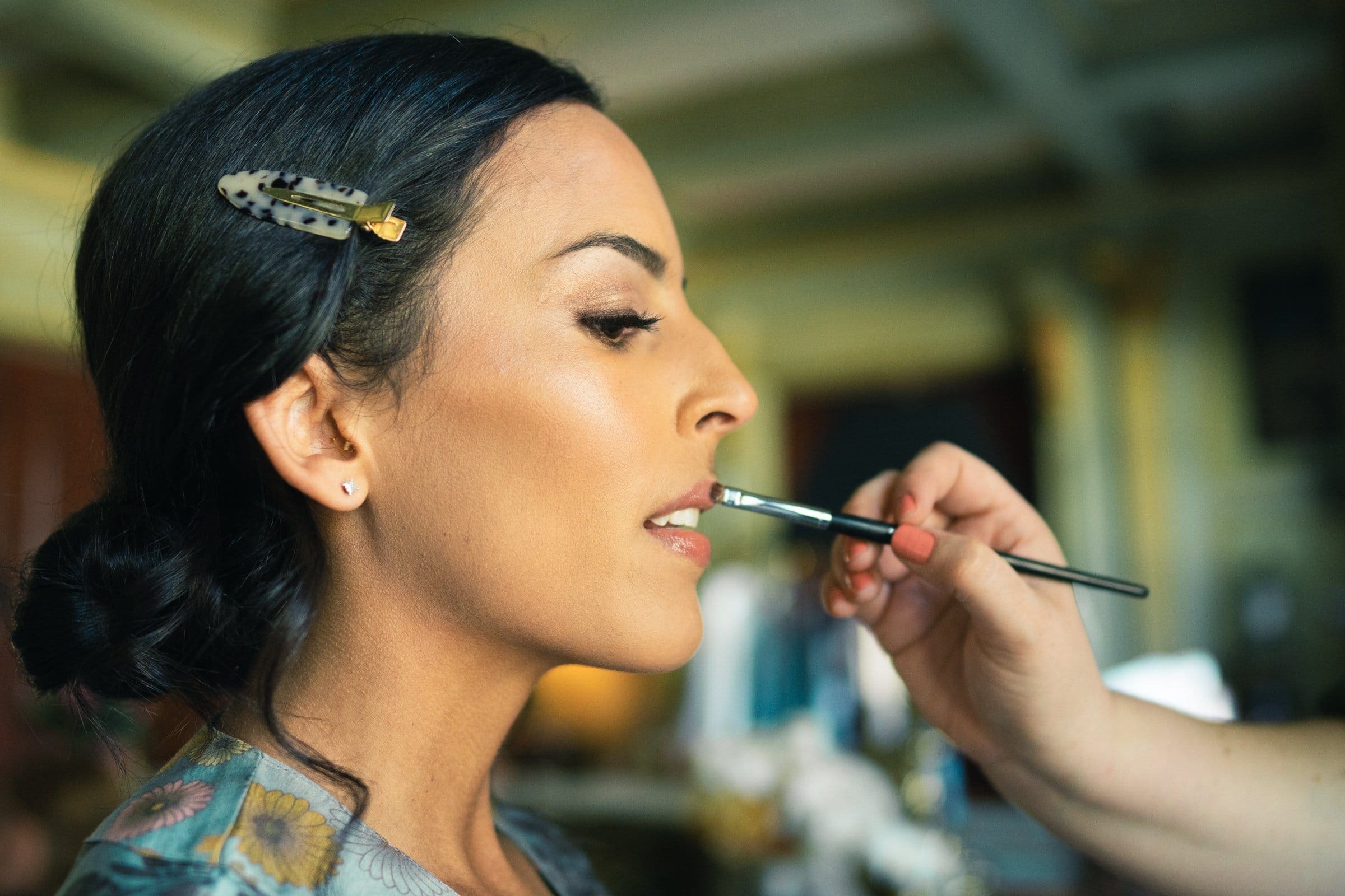 A woman is having makeup artfully applied by another person using a brush. She wears a hair clip, sitting in a well-lit room that perfectly captures her profile and the intricate makeup process, reminiscent of a Raleigh photographer's attention to detail.