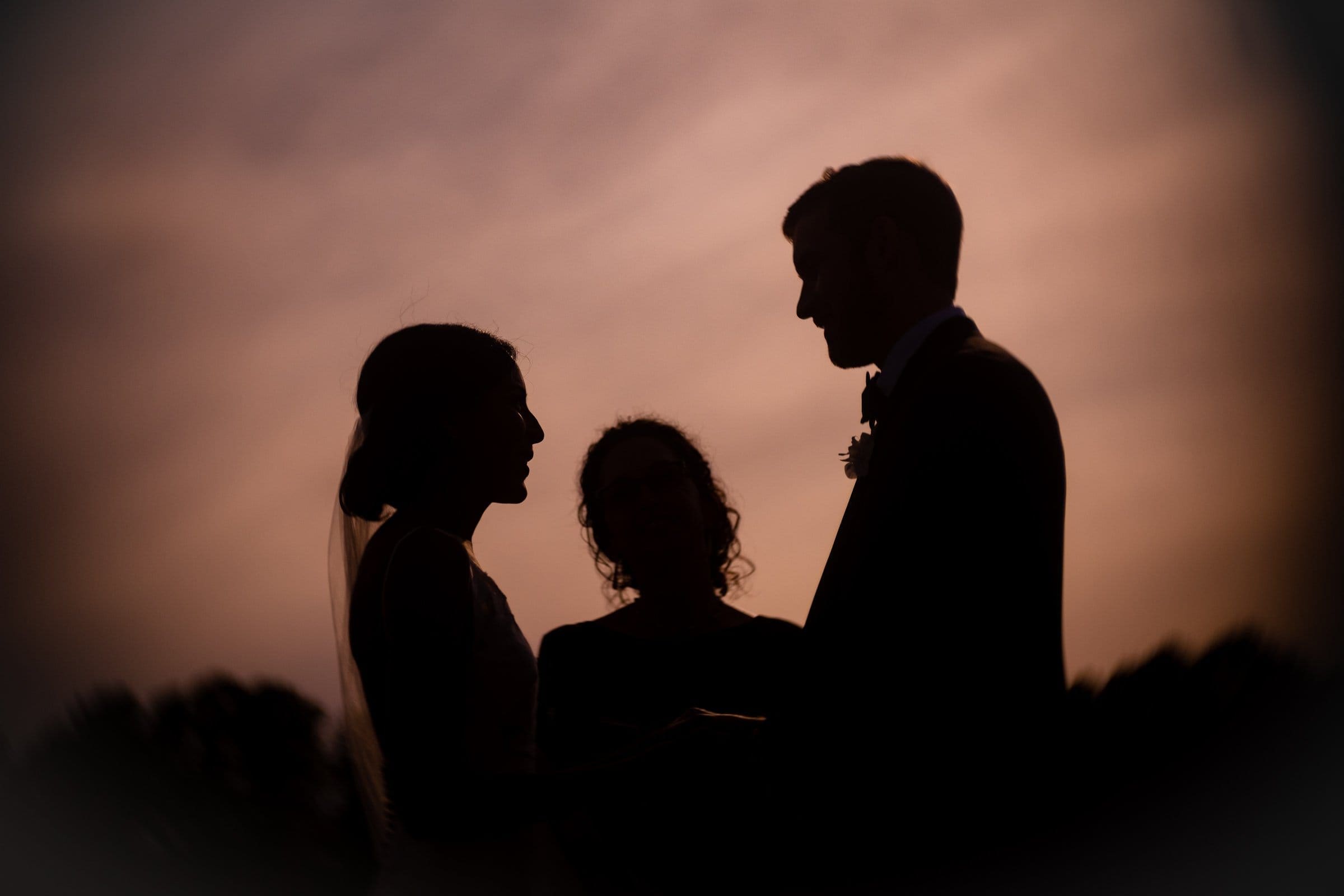 Silhouetted image of a couple exchanging vows outdoors at sunset, captured by a Raleigh photographer. The sky is a soft blend of orange and purple hues, creating a romantic atmosphere as an officiant stands by.