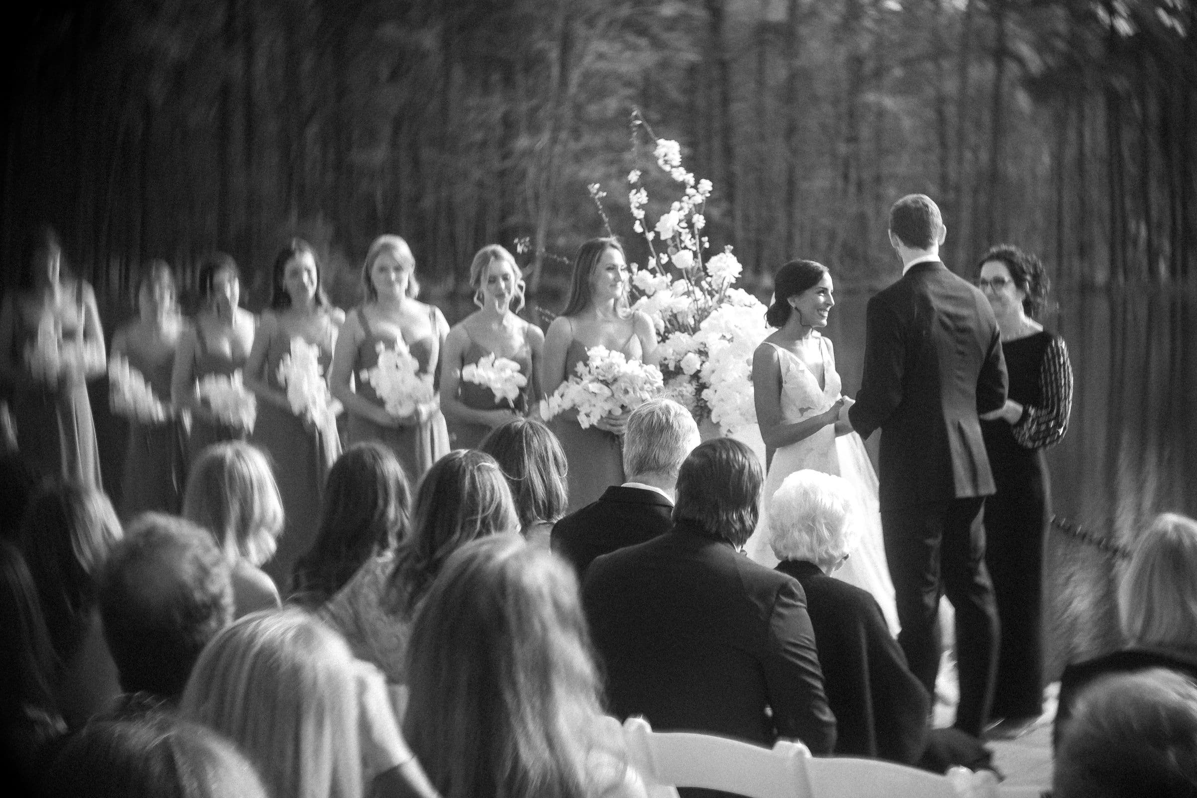 Black and white photo of a wedding ceremony captured by a Raleigh photographer. The bride and groom stand facing each other, surrounded by bridesmaids holding flower bouquets. Guests are seated, watching the couple, while trees create a serene backdrop.