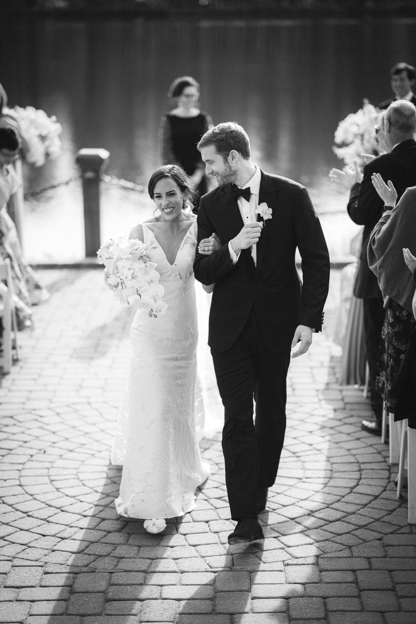 A bride and groom walk arm in arm down a cobblestone aisle, smiling joyfully as a Raleigh photographer captures the moment. The bride holds a bouquet of white flowers, and the groom sports a suit with a boutonniere. Guests stand and clap around them, with a shimmering body of water in the background.