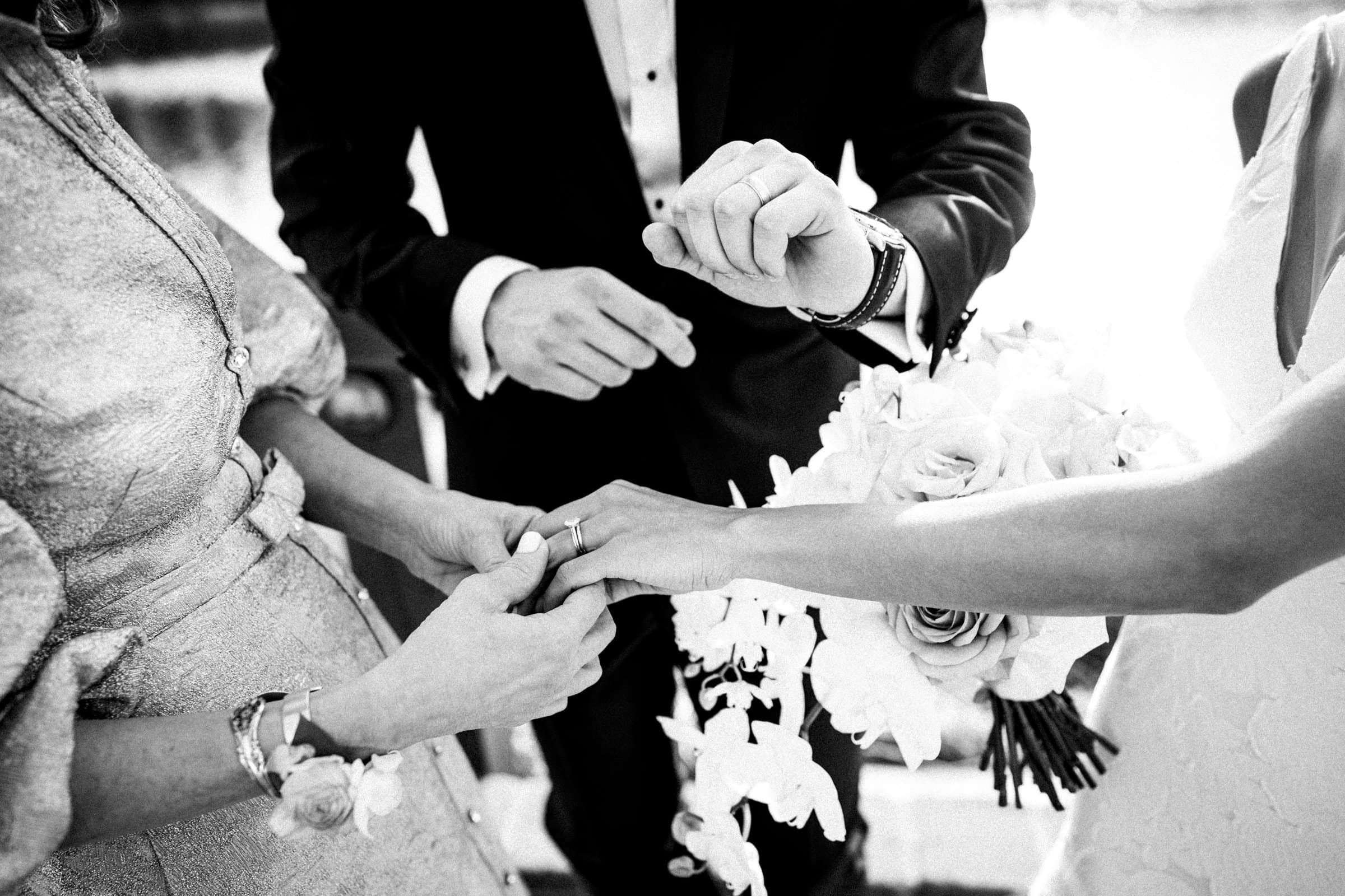 A black and white image captured by a Raleigh photographer portrays a wedding ceremony. A person is placing a ring on the bride's finger while she holds a bouquet of flowers. The groom, in his suit, stands beside them, with focus on their hands and the delicate action of the moment.