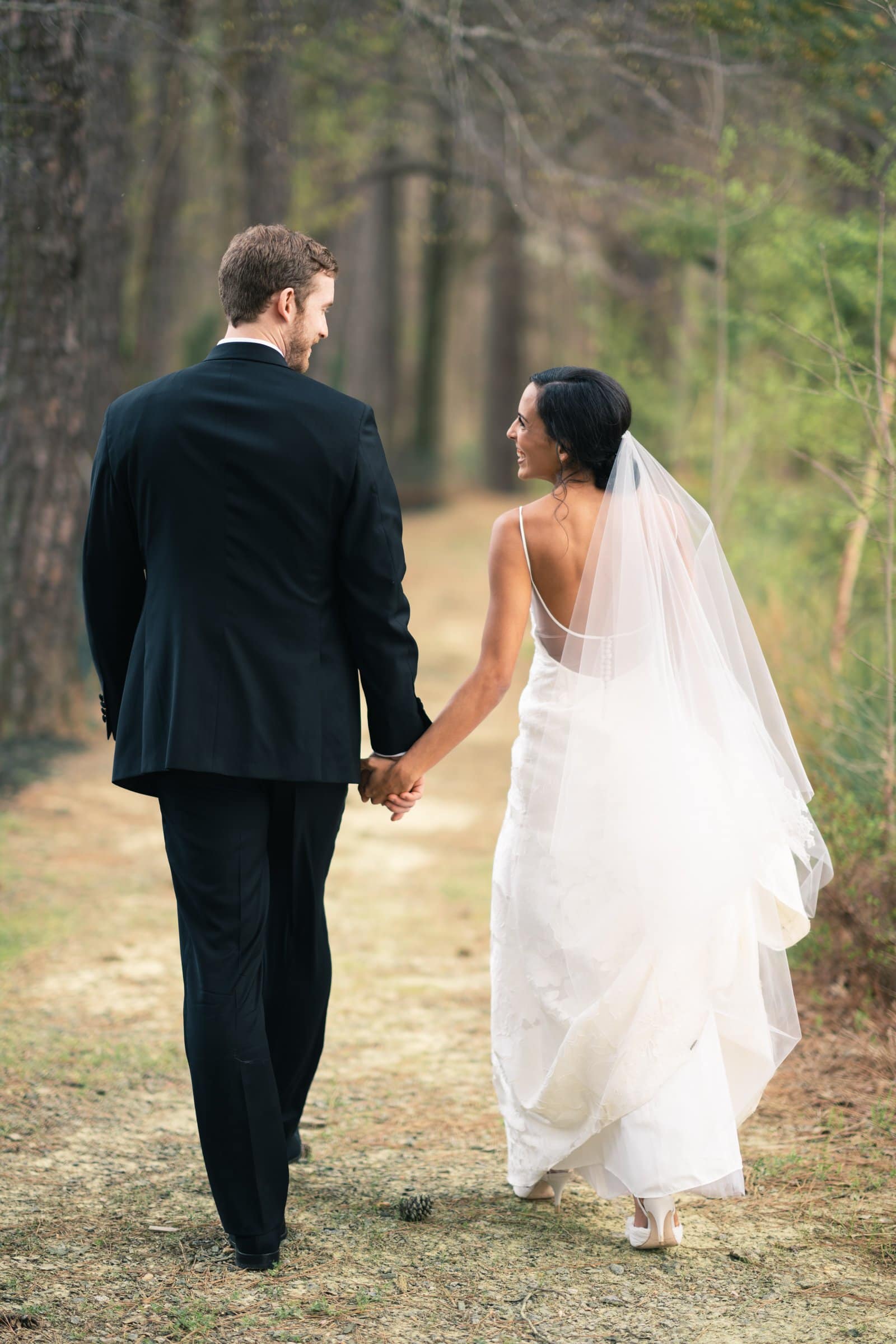 A bride and groom walk hand in hand down a wooded path, captured beautifully by a Raleigh photographer. The groom wears a black suit, and the bride is in a white dress with a veil. They look at each other affectionately, surrounded by tall trees and lush greenery.