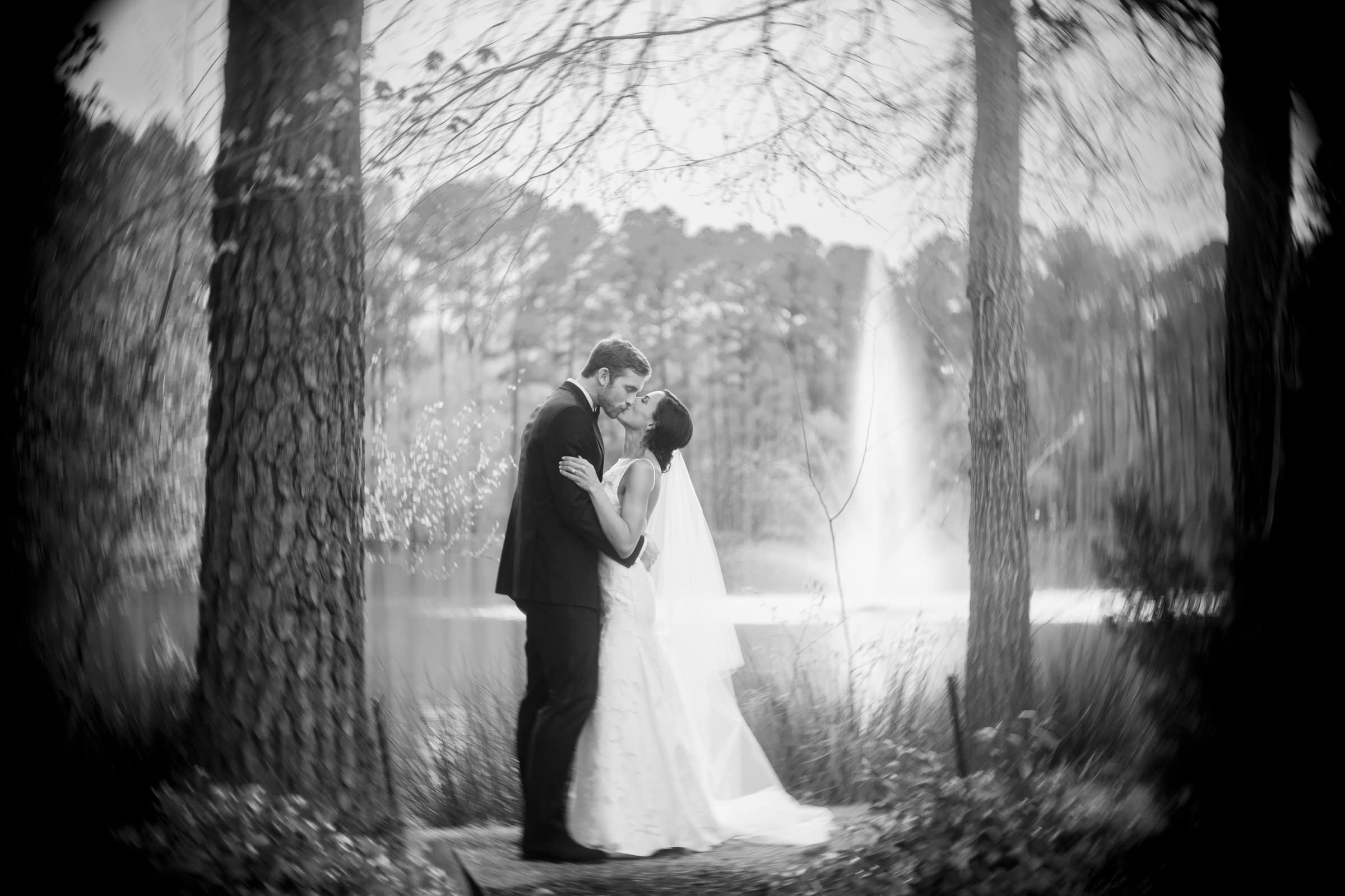 A bride and groom share a kiss in a serene forest setting, beautifully captured by a Raleigh photographer. They stand between two tall trees with a fountain visible in the background. The bride wears a veil, and the scene has a soft, dreamy quality.