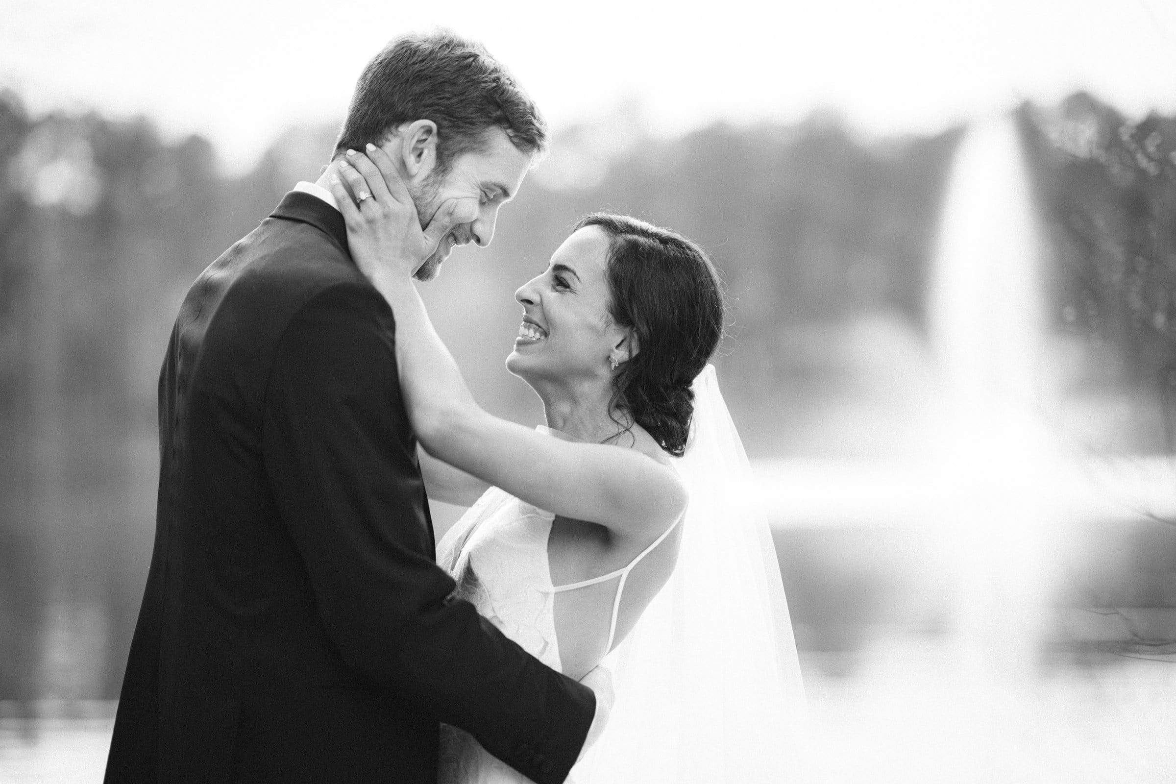 A black and white photo, skillfully captured by a Raleigh photographer, reveals a bride and groom in wedding attire, smiling at each other. The bride, wearing a veil, gently holds the groom's face as they stand before a blurred background of water and a fountain.