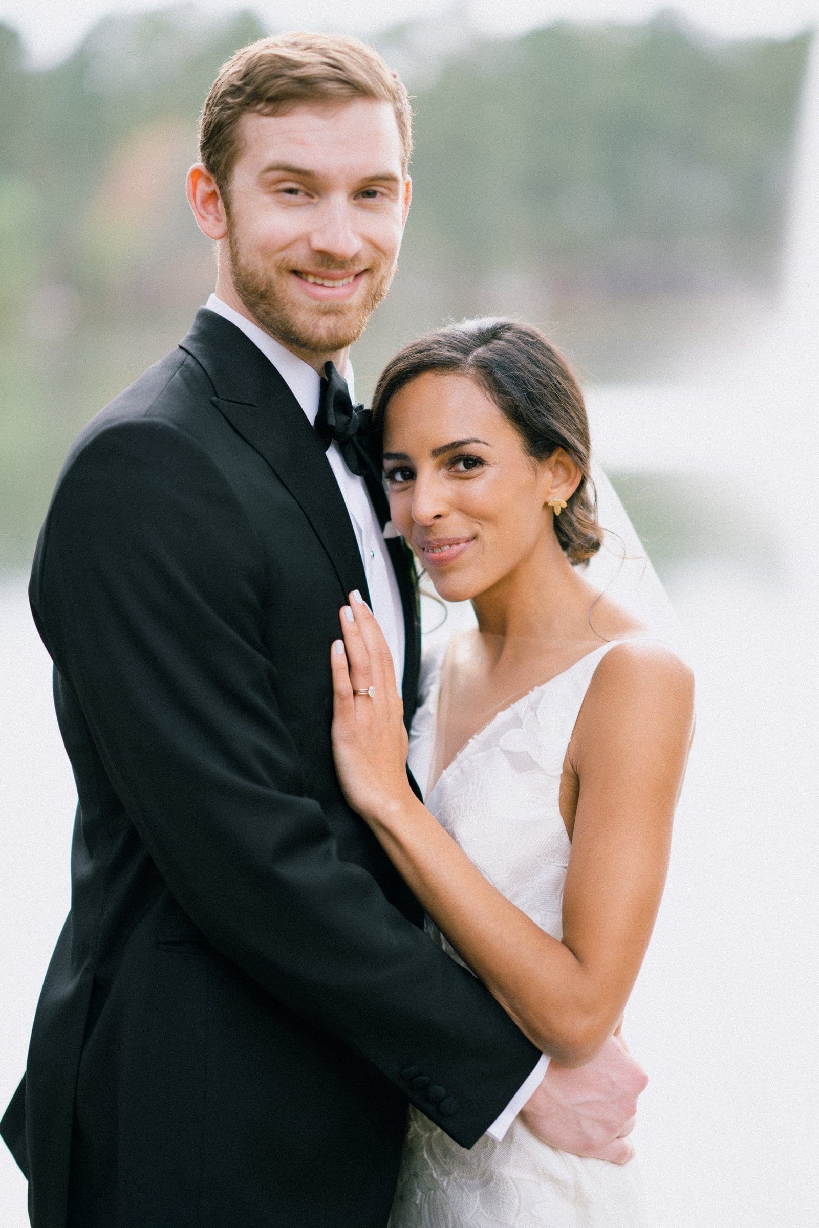 A bride and groom smile while embracing, captured beautifully by a Raleigh photographer. The bride is wearing a white dress and veil, and the groom is in a black tuxedo. They are outdoors, with a blurred background of greenery and water enhancing their joy.
