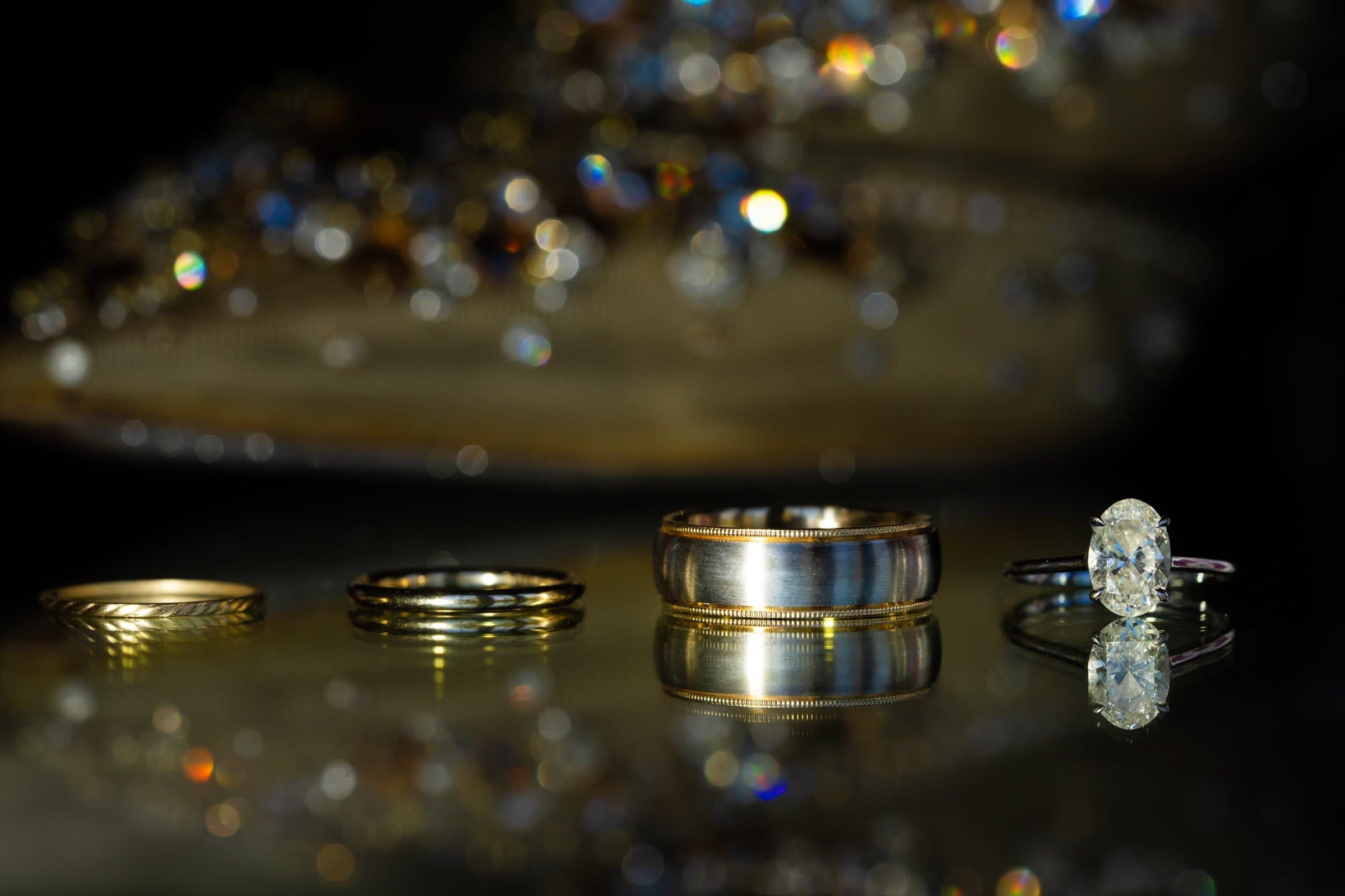 Several rings are displayed on a reflective surface captured by a skilled Raleigh photographer. From left to right: two gold bands, a wider metallic band with a gold rim, and a silver ring with an oval diamond. Sparkling bokeh lights twinkle in the blurred background.
