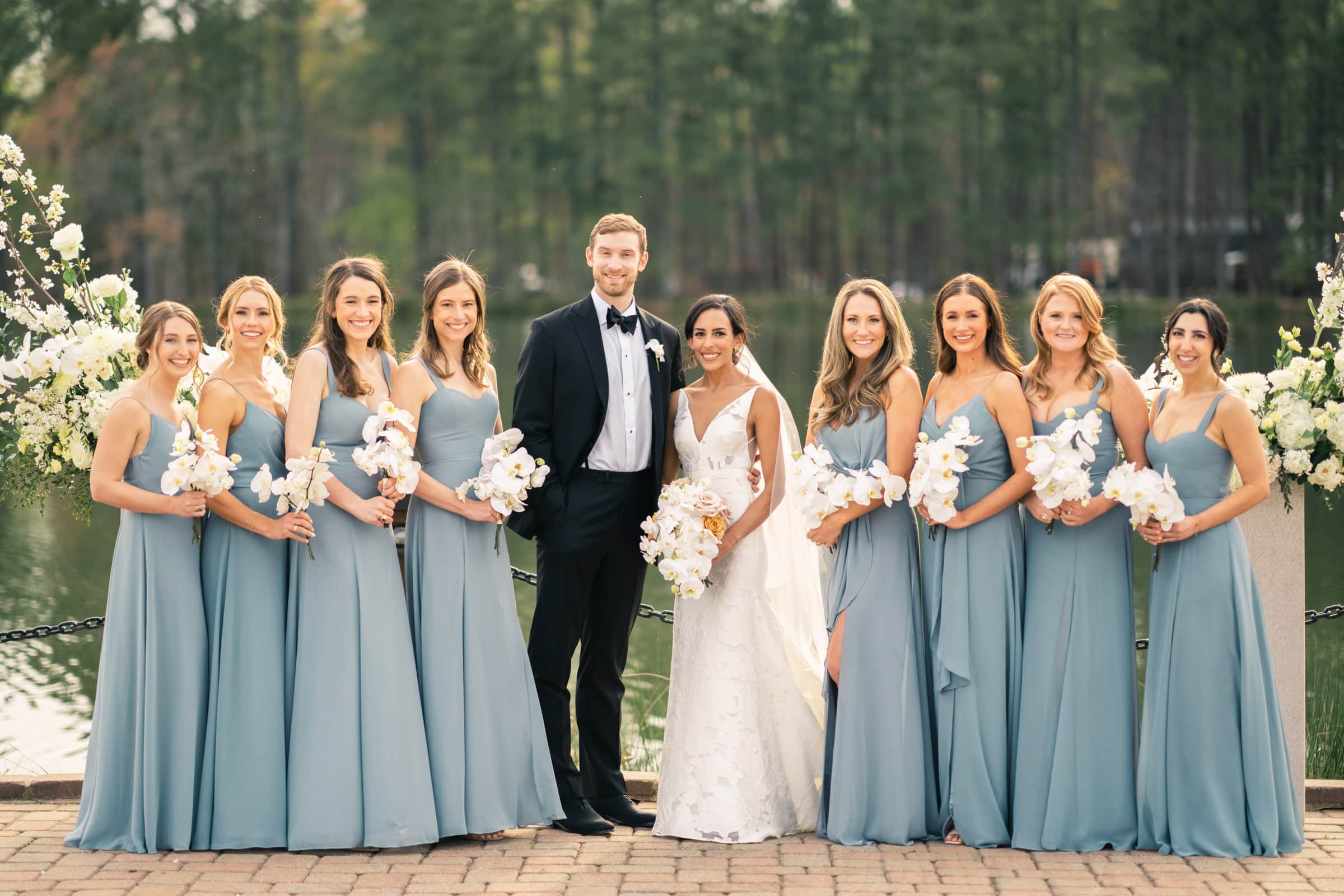 A Raleigh photographer captures a bride in a white gown and groom in a black tuxedo, joined by seven bridesmaids in light blue dresses. They stand outdoors by a picturesque lake, surrounded by trees, all holding elegant white flower bouquets.