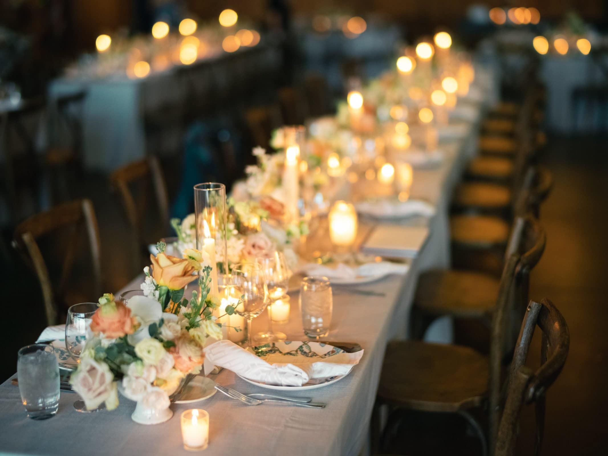 A beautifully set long dinner table with elegant floral arrangements and glowing candles, captured by a talented Raleigh photographer. Wooden chairs line the table, adorned with white linen, glassware, and folded napkins, creating a warm and inviting atmosphere.