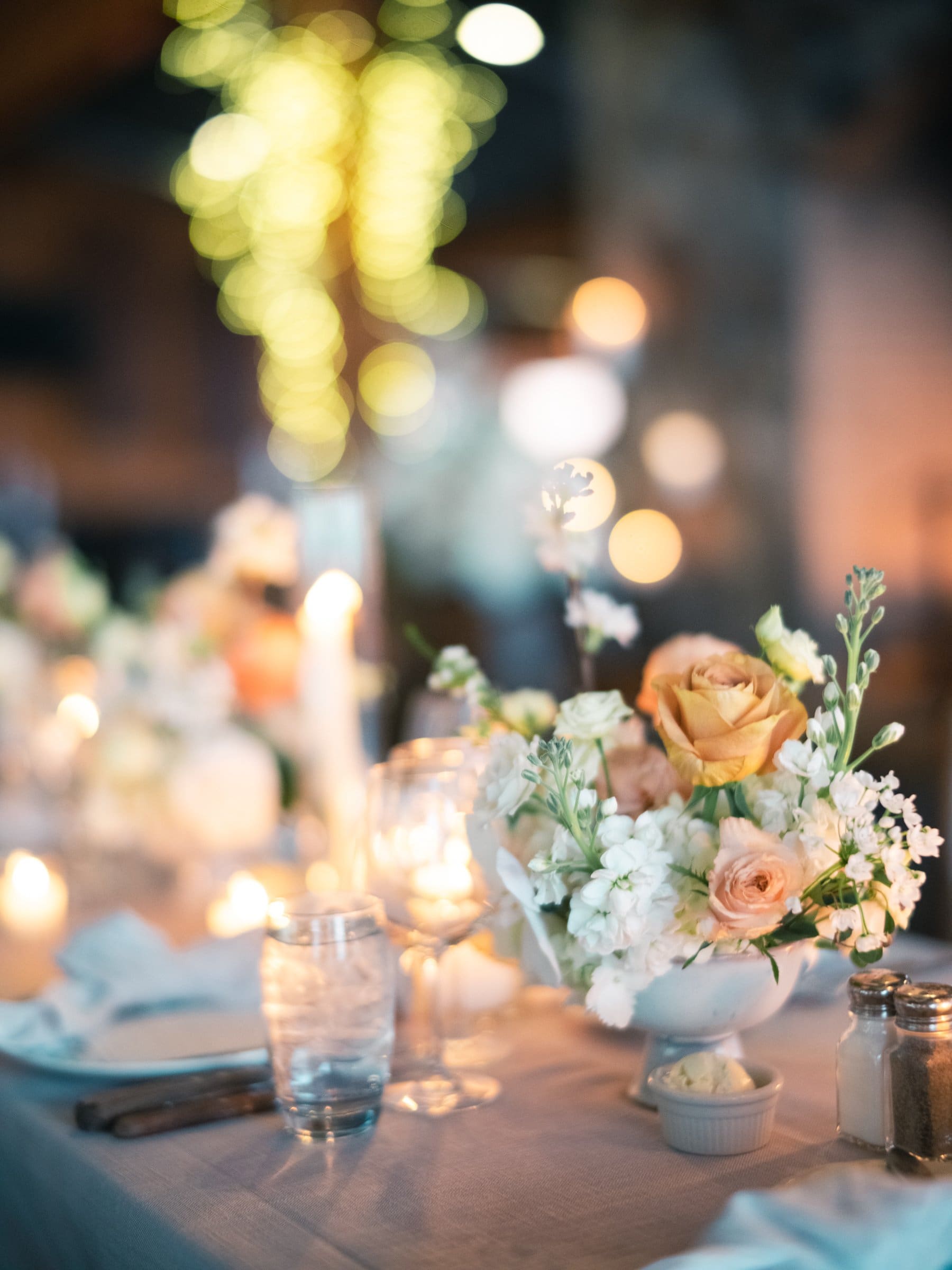 A beautifully set table with floral centerpieces featuring roses and white blooms, captured by a Raleigh photographer. Lit candles, glasses, and cutlery adorn the setting. The background is softly blurred with warm, glowing lights, creating an intimate ambiance.