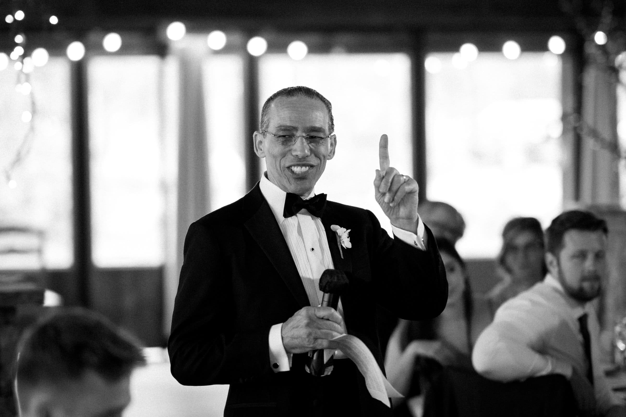 A man in a tuxedo is smiling and holding a microphone while gesturing upward, caught brilliantly by a Raleigh photographer. He appears to be giving a speech at a formal event, with string lights glistening above and several seated people in the background.