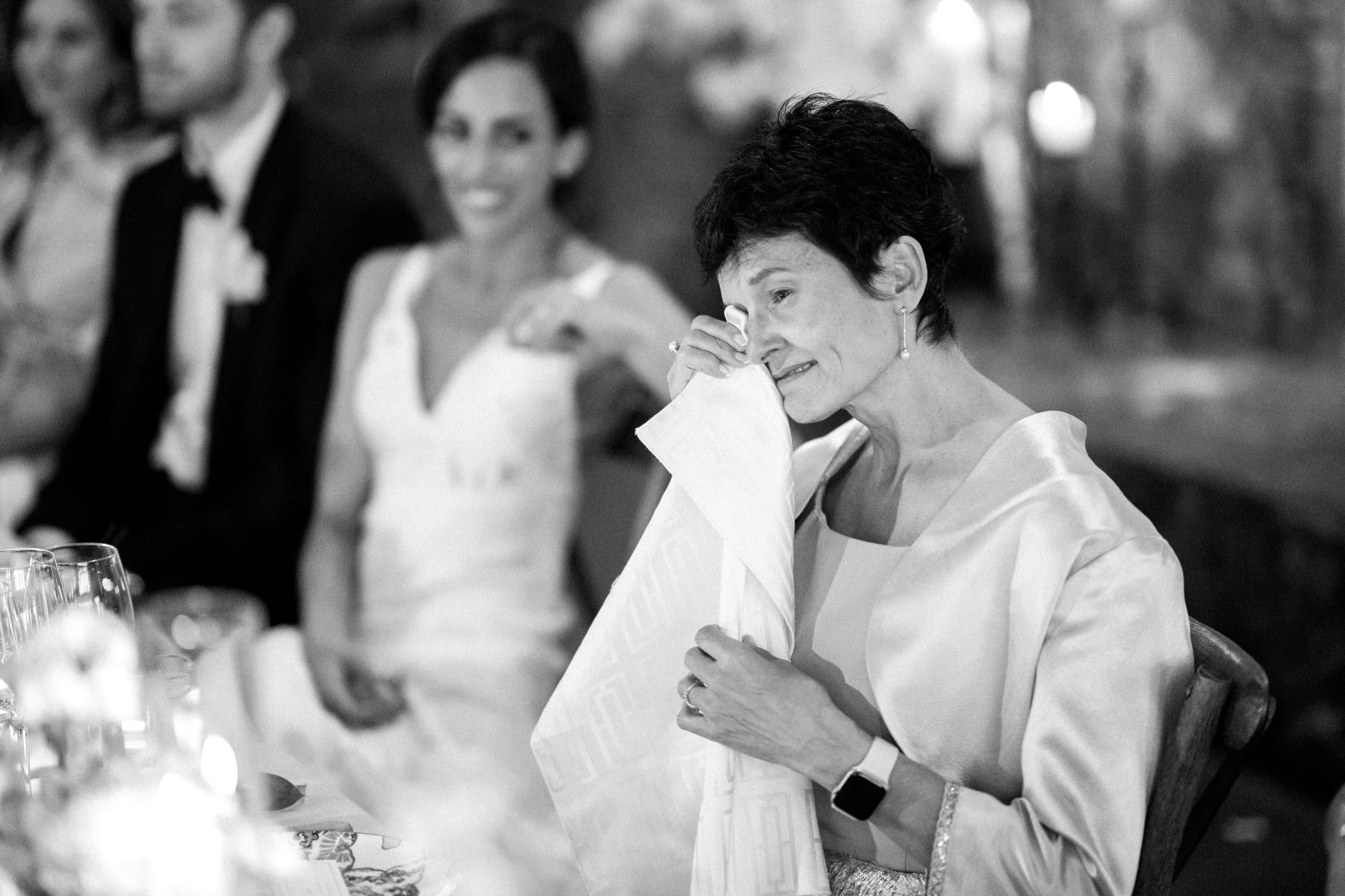A woman in a satin shawl, seated at a table, wipes away tears with a napkin, her smile soft and genuine. Captured by a Raleigh photographer, the scene features the bride, groom, and another guest in the background, all elegantly dressed in this timeless black-and-white image.