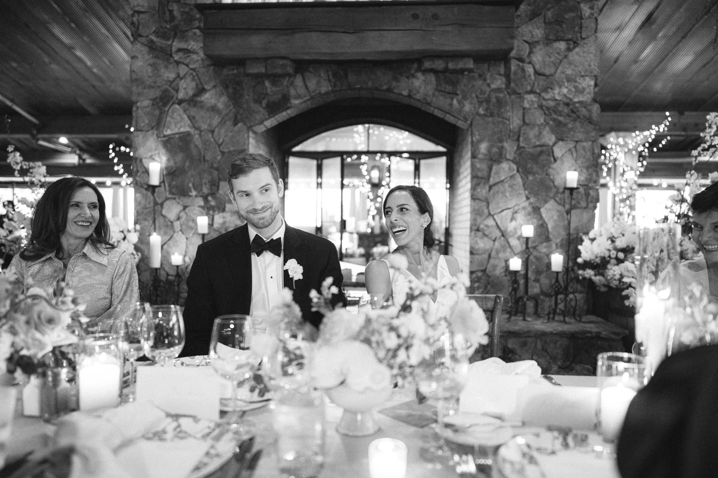 Captured by a Raleigh photographer, this black and white photo showcases a wedding reception. The groom in a tuxedo sits beside the bride in a white dress, both smiling amid guests at a table with candles, flowers, and glasses, all set against a rustic stone backdrop.