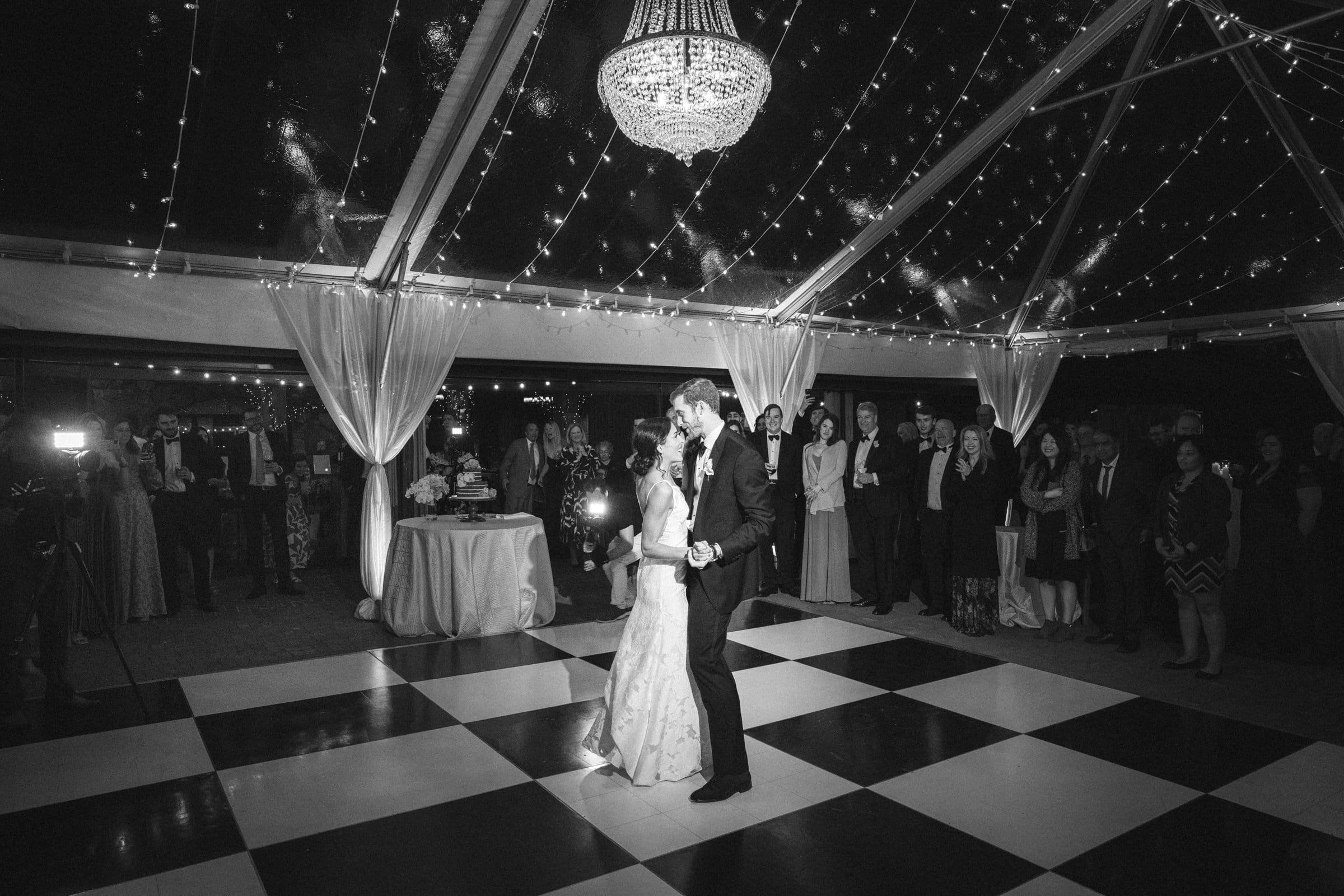 A bride and groom share a first dance on a checkered dance floor under a chandelier and string lights, beautifully captured by a Raleigh photographer. Guests stand around them, watching the moment in the dimly lit, elegant venue.