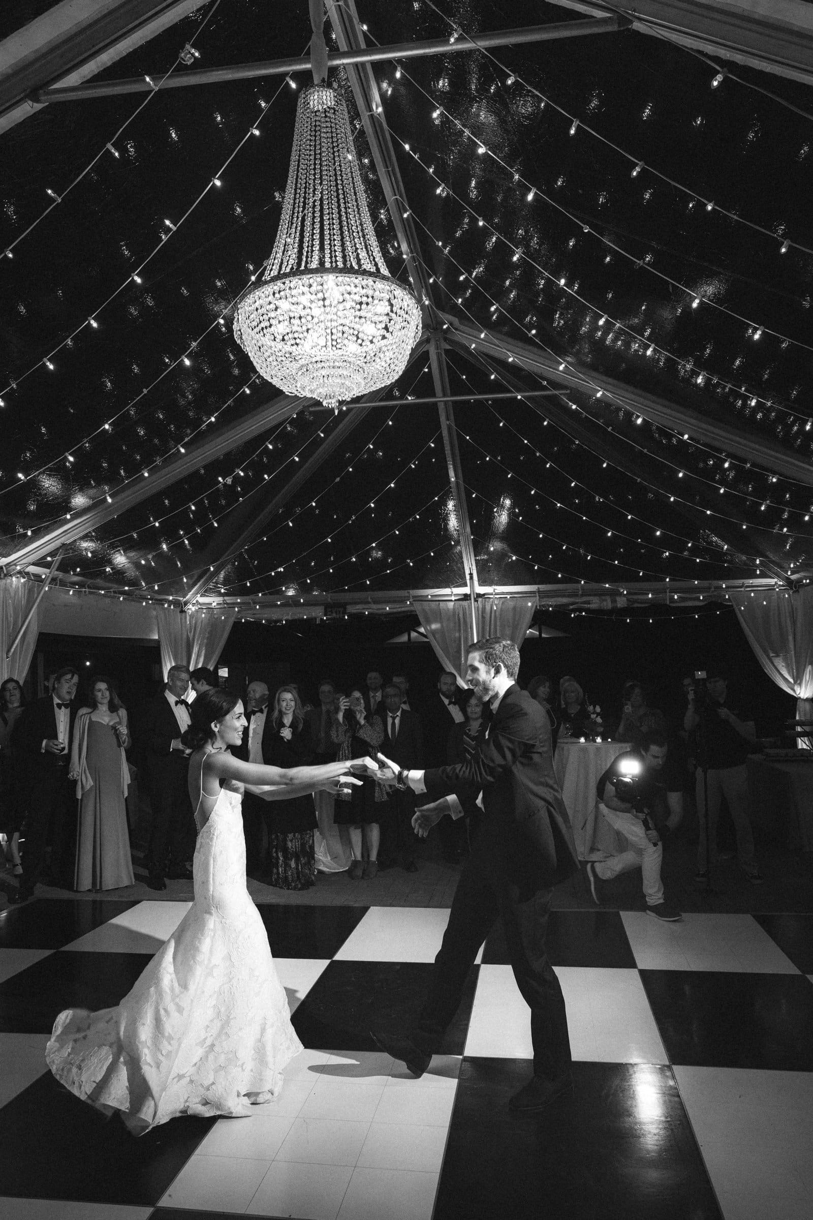 Under a sparkling chandelier in a tent adorned with string lights, captured by a Raleigh photographer, the bride and groom dance gracefully. The couple is elegantly dressed, with the bride in a flowing gown and the groom in a suit, as guests watch in admiration.