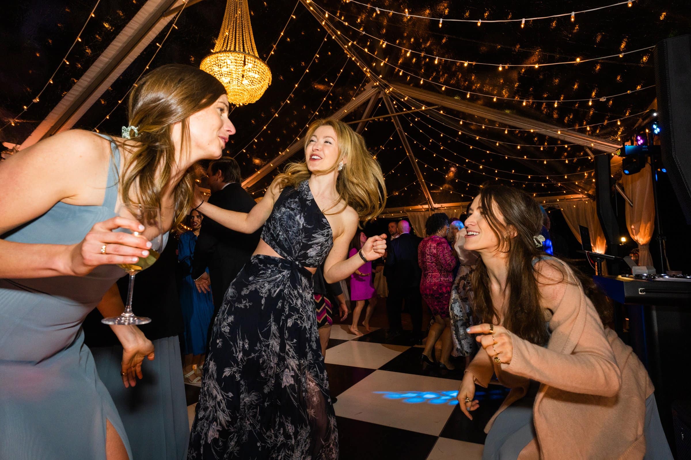 Three women in dresses laugh and dance under a canopy adorned with string lights and a chandelier, captured perfectly by a Raleigh photographer. The setting is festive, with a checkered dance floor and guests enjoying the lively atmosphere.