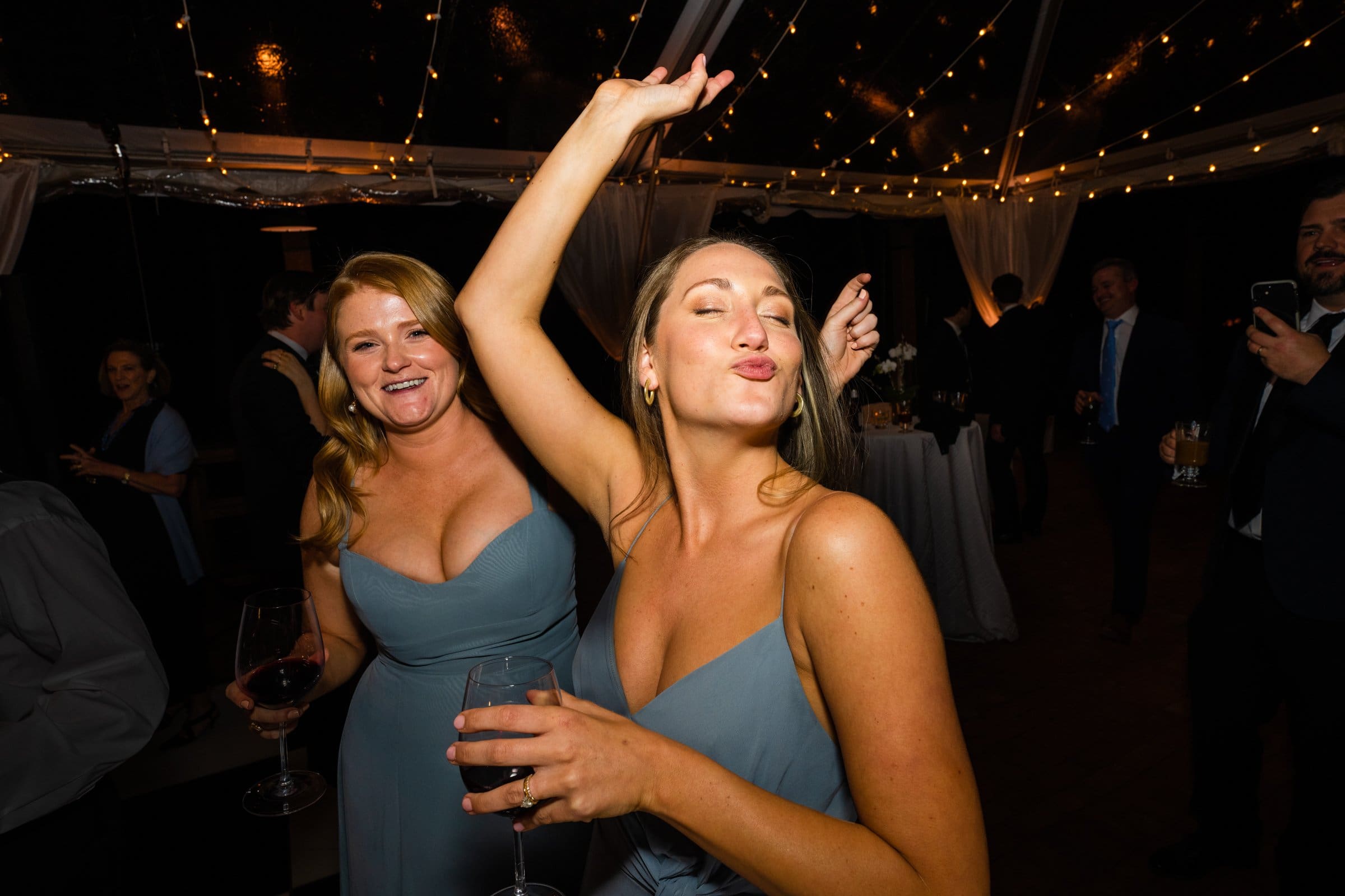 Two women in blue dresses joyfully dance at a party captured by a Raleigh photographer. One woman playfully closes her eyes and holds a drink, while string lights twinkle in the background. Other guests can be seen enjoying the vibrant event.