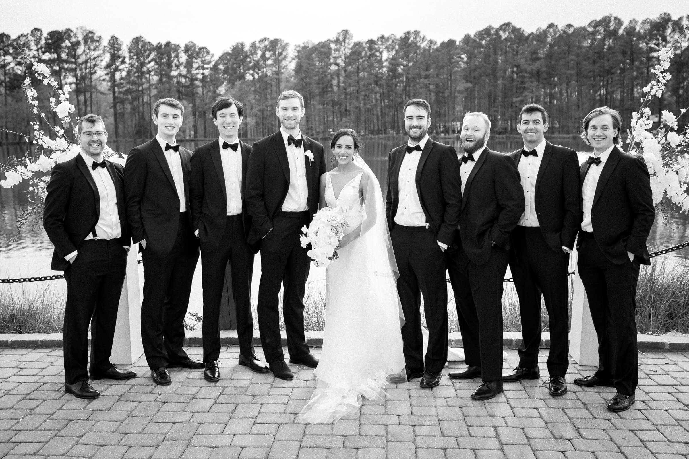 A bride in a white gown holds a bouquet, surrounded by eight groomsmen in tuxedos. Their smiles shine against the serene lake backdrop. Captured by a Raleigh photographer, this black-and-white scene exudes a classic and timeless feel.