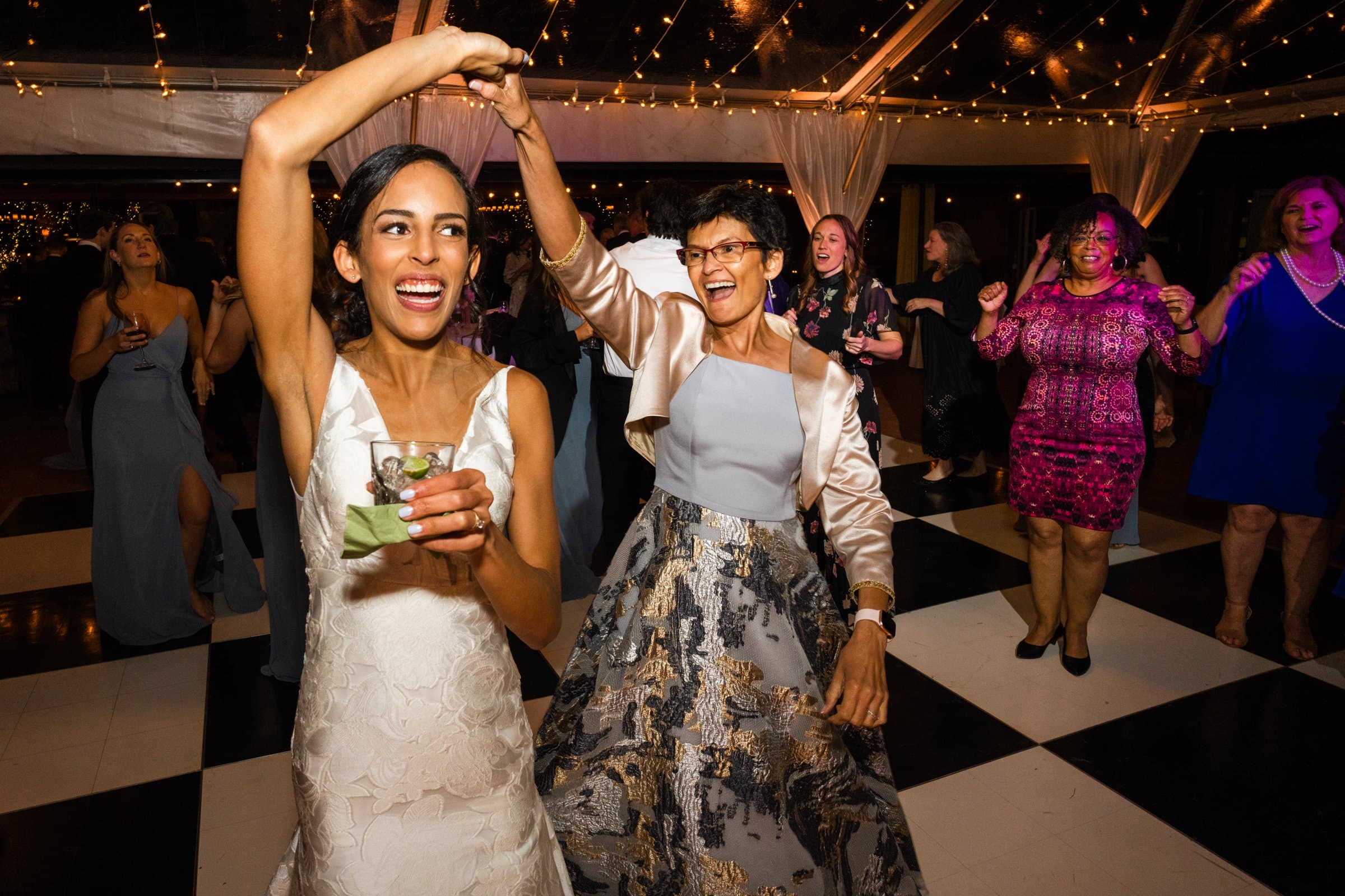 A joyful bride in a white dress dances with a woman in an elegant skirt and jacket at a lively party captured by a Raleigh photographer. They are surrounded by smiling guests on a checkerboard dance floor under string lights.