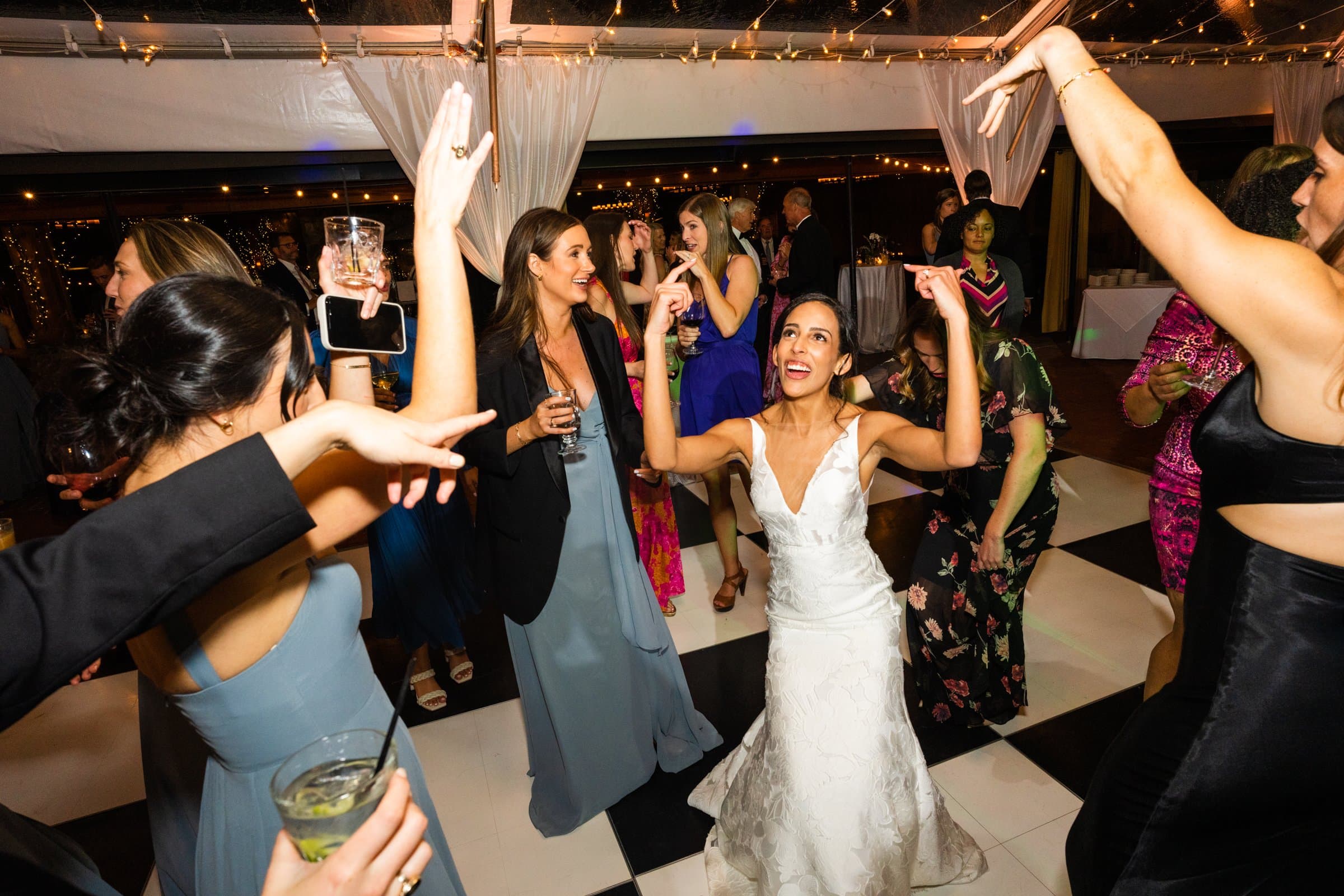 A bride in a white dress joyfully dances on a black and white checkered floor, captured vividly by the Raleigh photographer. String lights adorn the venue as guests in formal attire smile, hold drinks, and enjoy the lively, festive atmosphere.