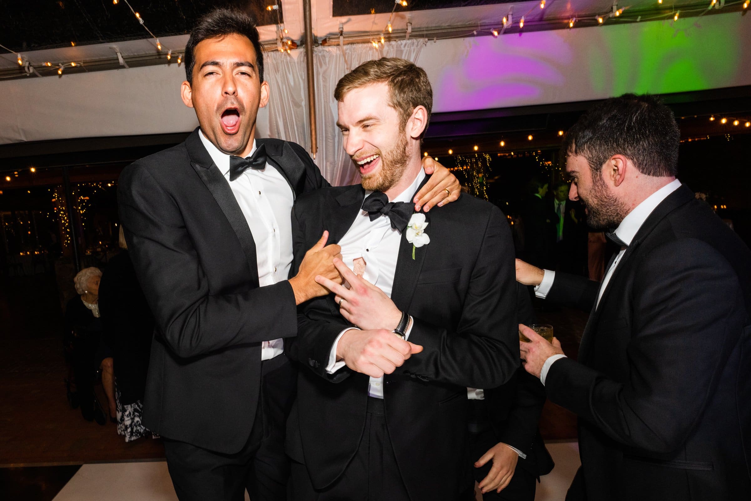 A group of four men in tuxedos are celebrating indoors, captured by a talented Raleigh photographer. Two men in the center are laughing and playfully interacting, while the other two are smiling. The background is decorated with lights, creating a festive atmosphere.